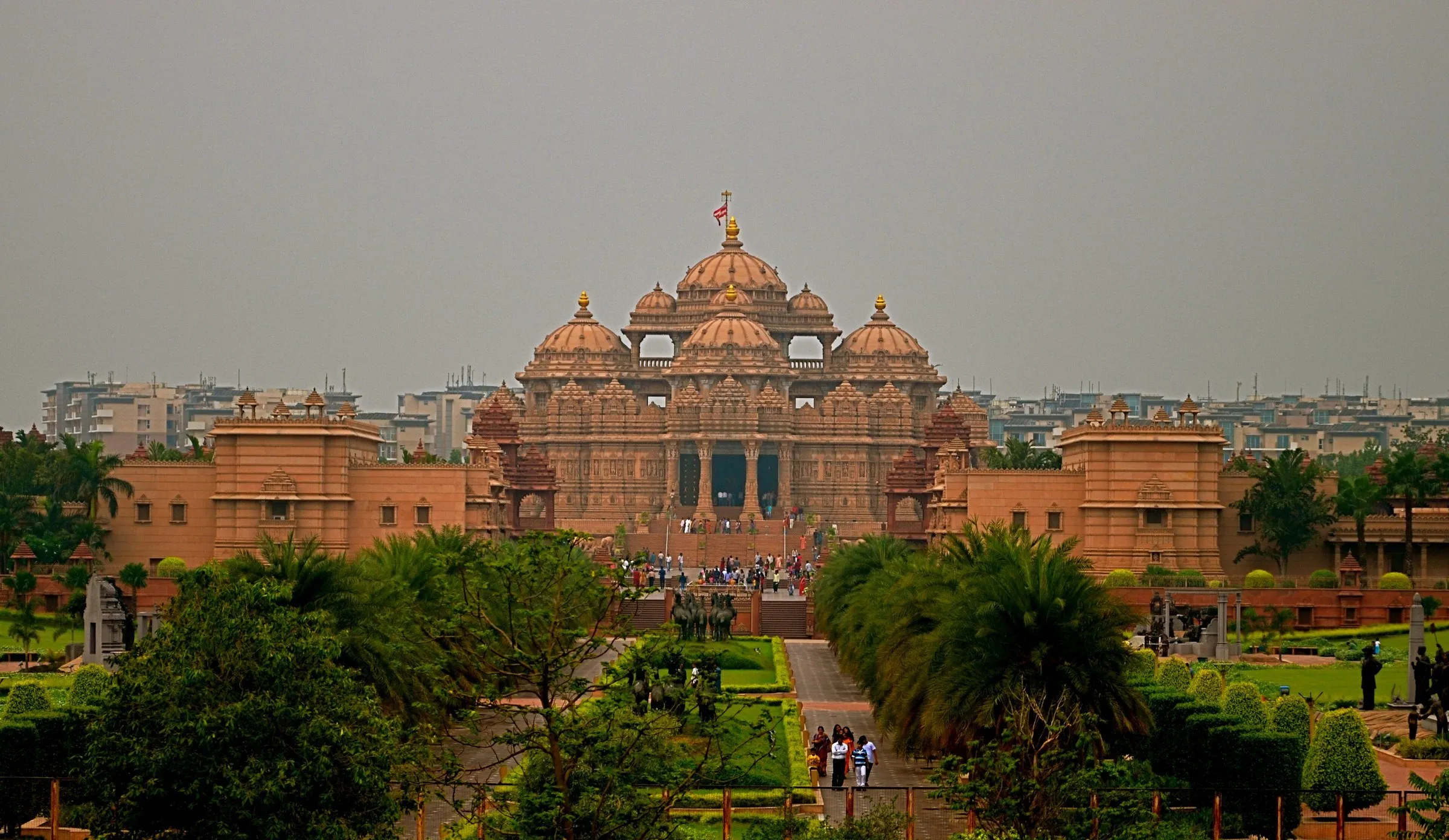 Fachada de arenito rosa do templo Akshardham com cúpulas e pináculos, vista de um jardim com palmeiras e visitantes pequenos.