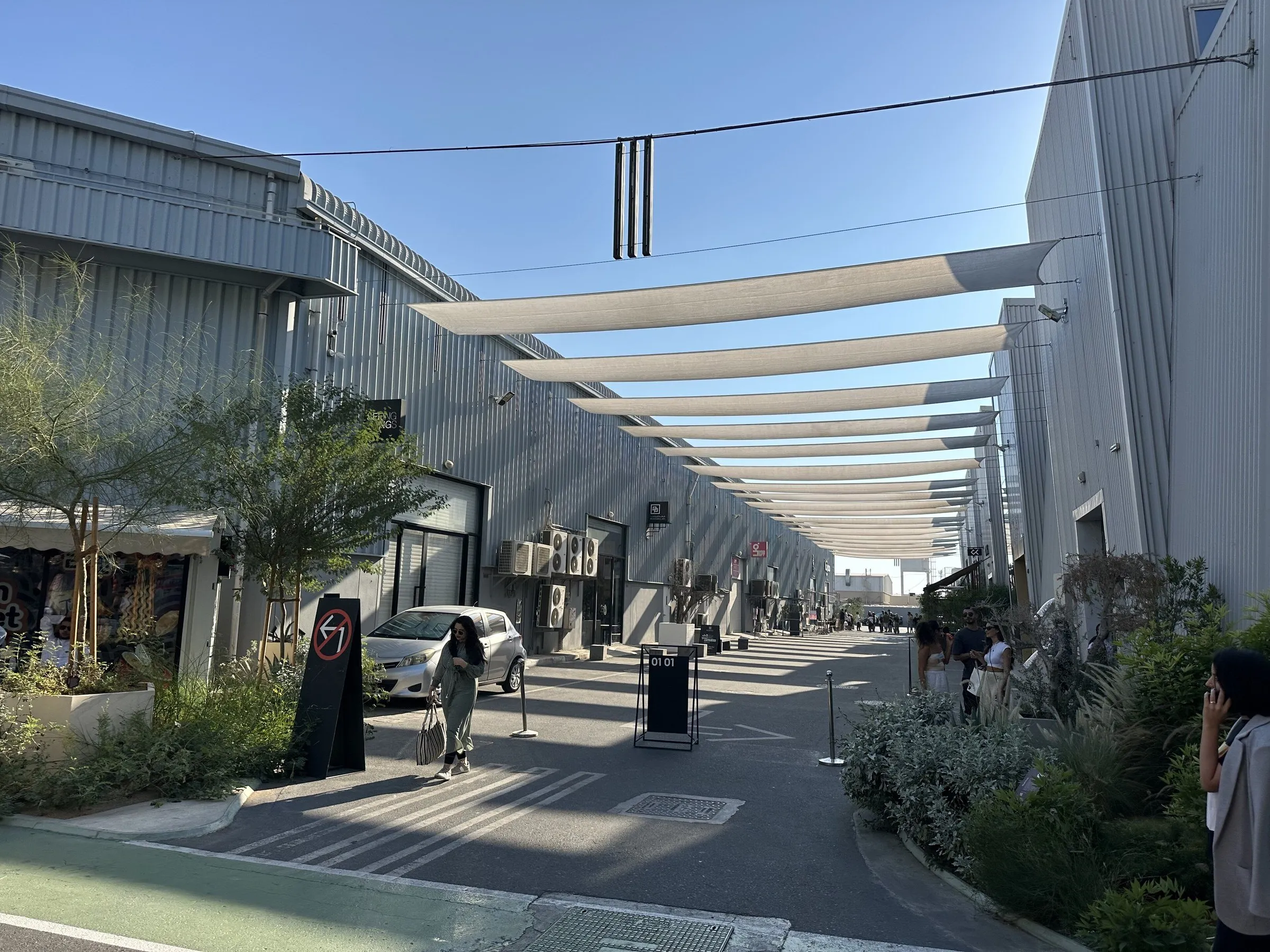 Rua entre galpões de metal da Alserkal Avenue, em Dubai, coberta por velas de sombra brancas, com plantas e poucas pessoas.