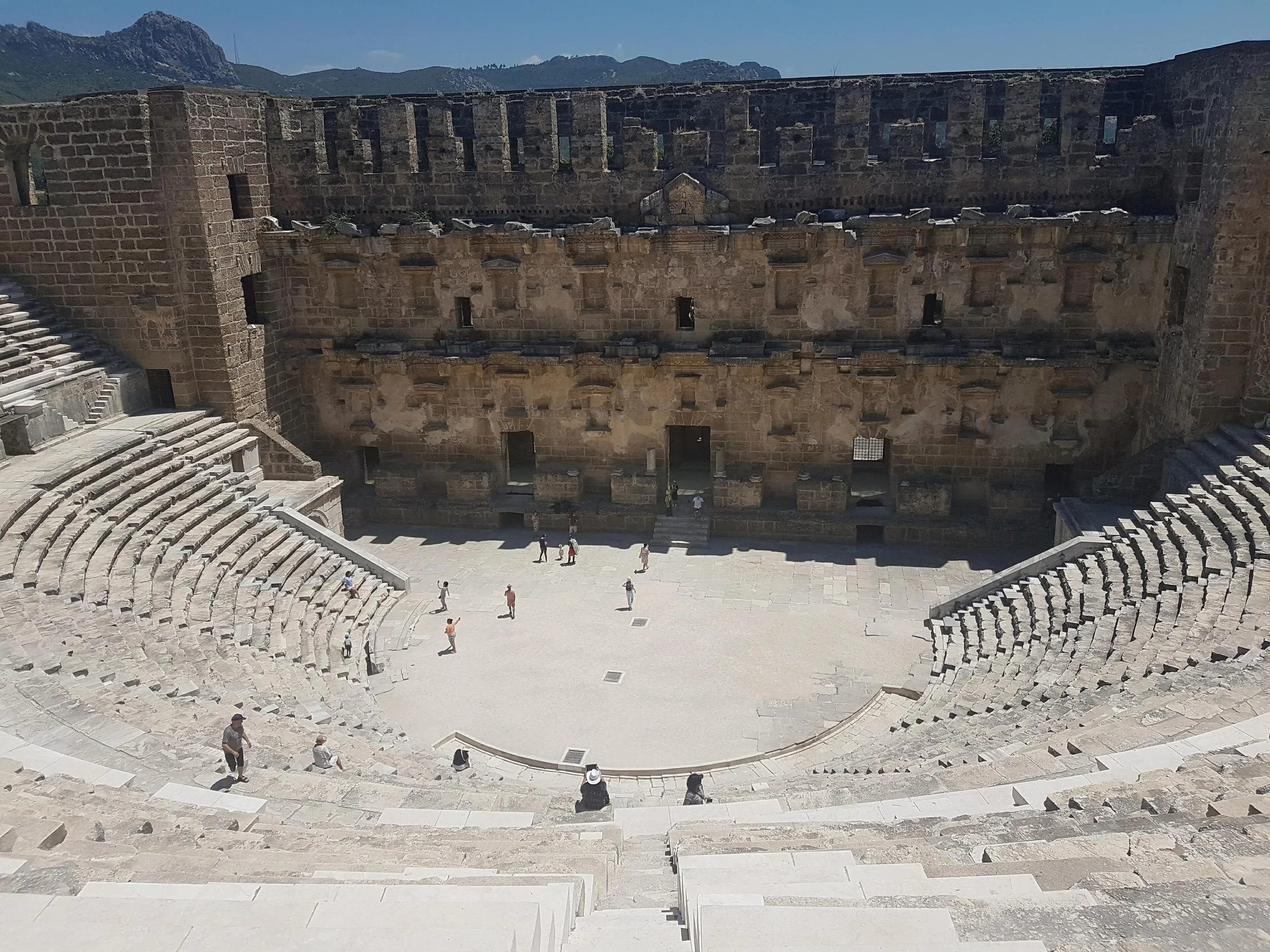 Teatro romano de Aspendos visto do alto da arquibancada, com a parede do palco quase inteira e poucos visitantes na arena.