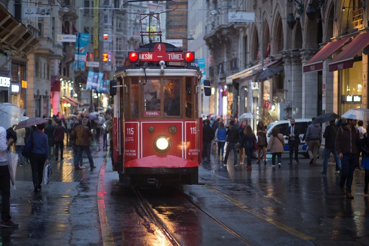 Bonde vermelho antigo Taksim–Tünel na Avenida İstiklal, com pedestres de guarda-chuva e calçamento molhado.