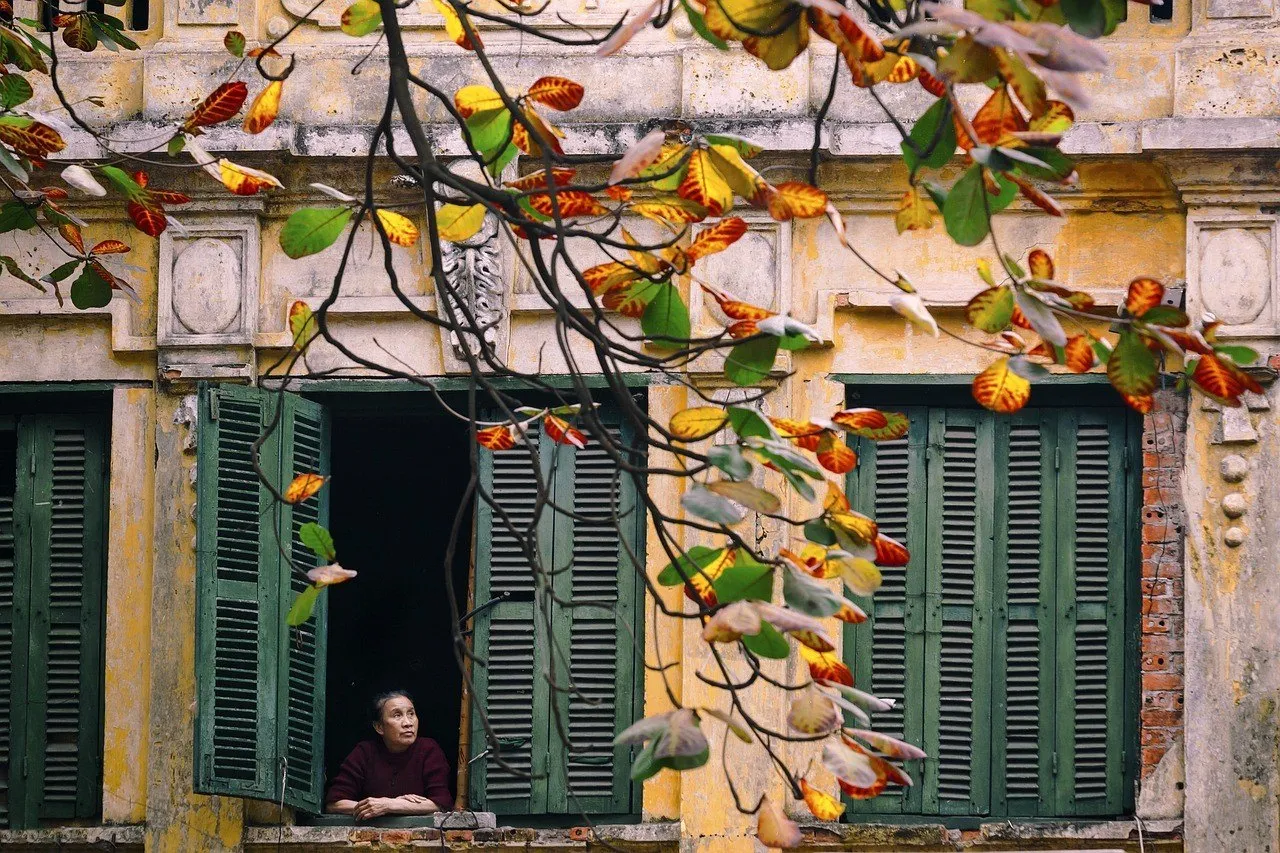 Fachada amarela com janelas verdes e galhos coloridos no Bairro Antigo, com uma moradora junto à janela.