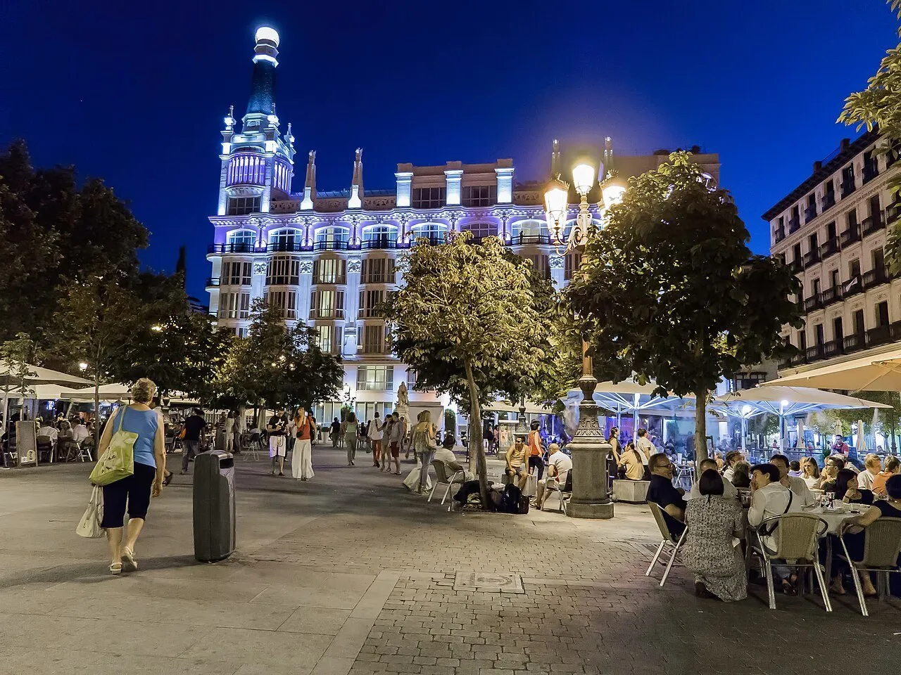 Plaza de Santa Ana à noite, com edifício iluminado, árvores, mesas de restaurantes e pedestres.