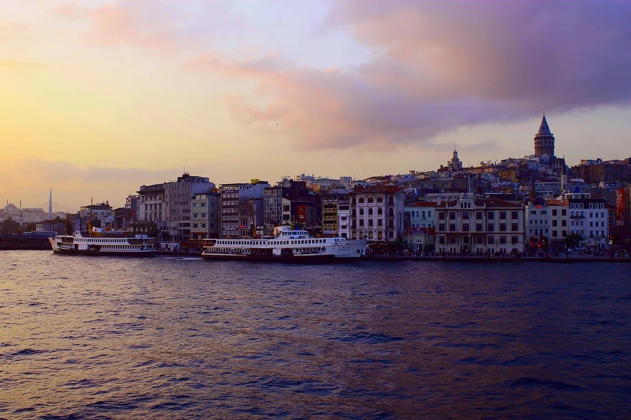 Balsas de passageiros na água ao entardecer, em frente ao casario de Karaköy, com a Torre de Gálata ao fundo e céu arroxeado.
