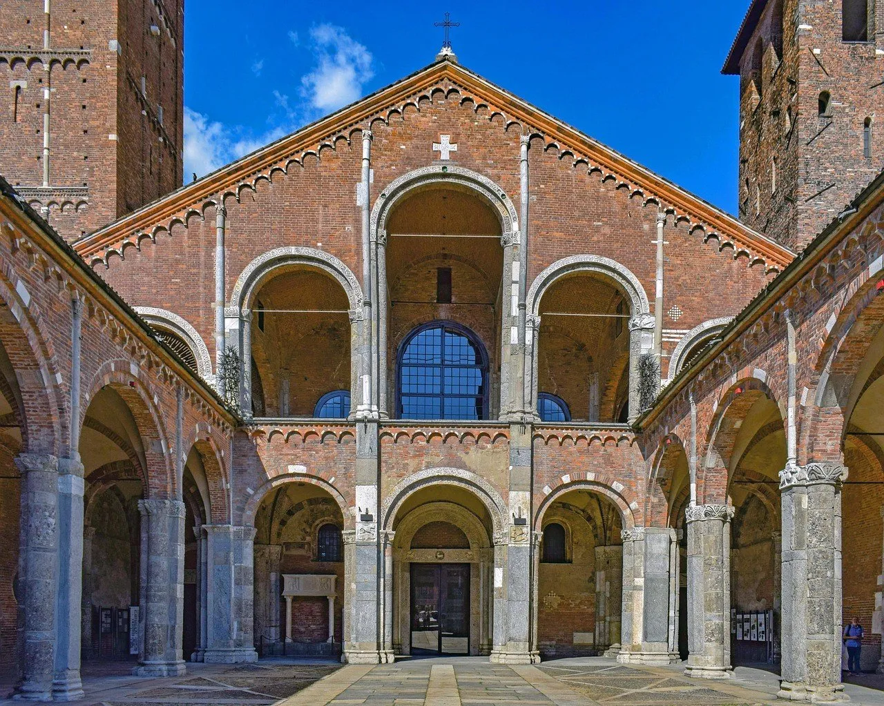 Fachada de tijolos da Basílica de Sant’Ambrogio vista do pátio entre arcadas, sob céu azul.
