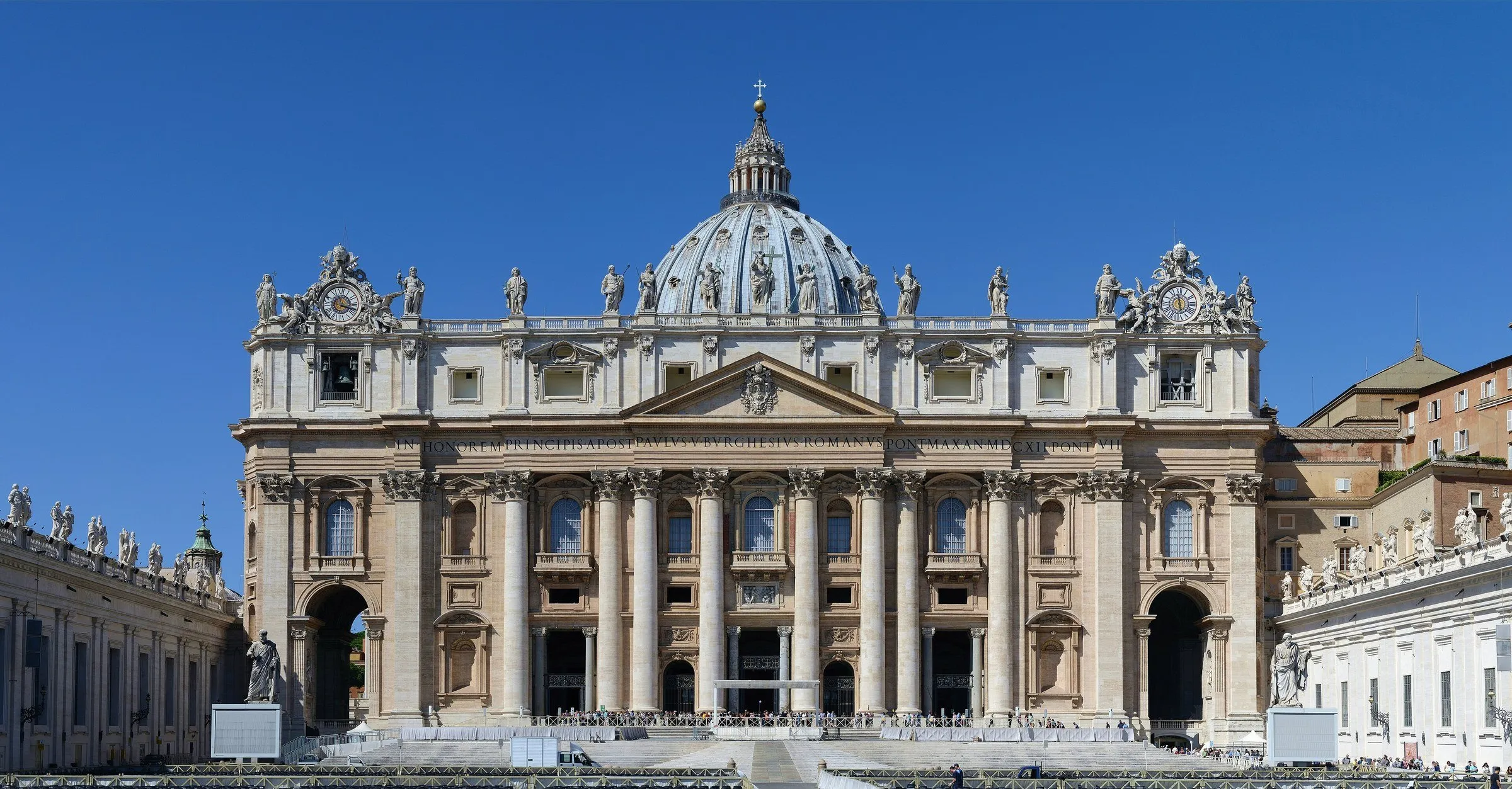 Fachada da Basílica de São Pedro, no Vaticano, com a cúpula ao centro e céu azul sem nuvens.