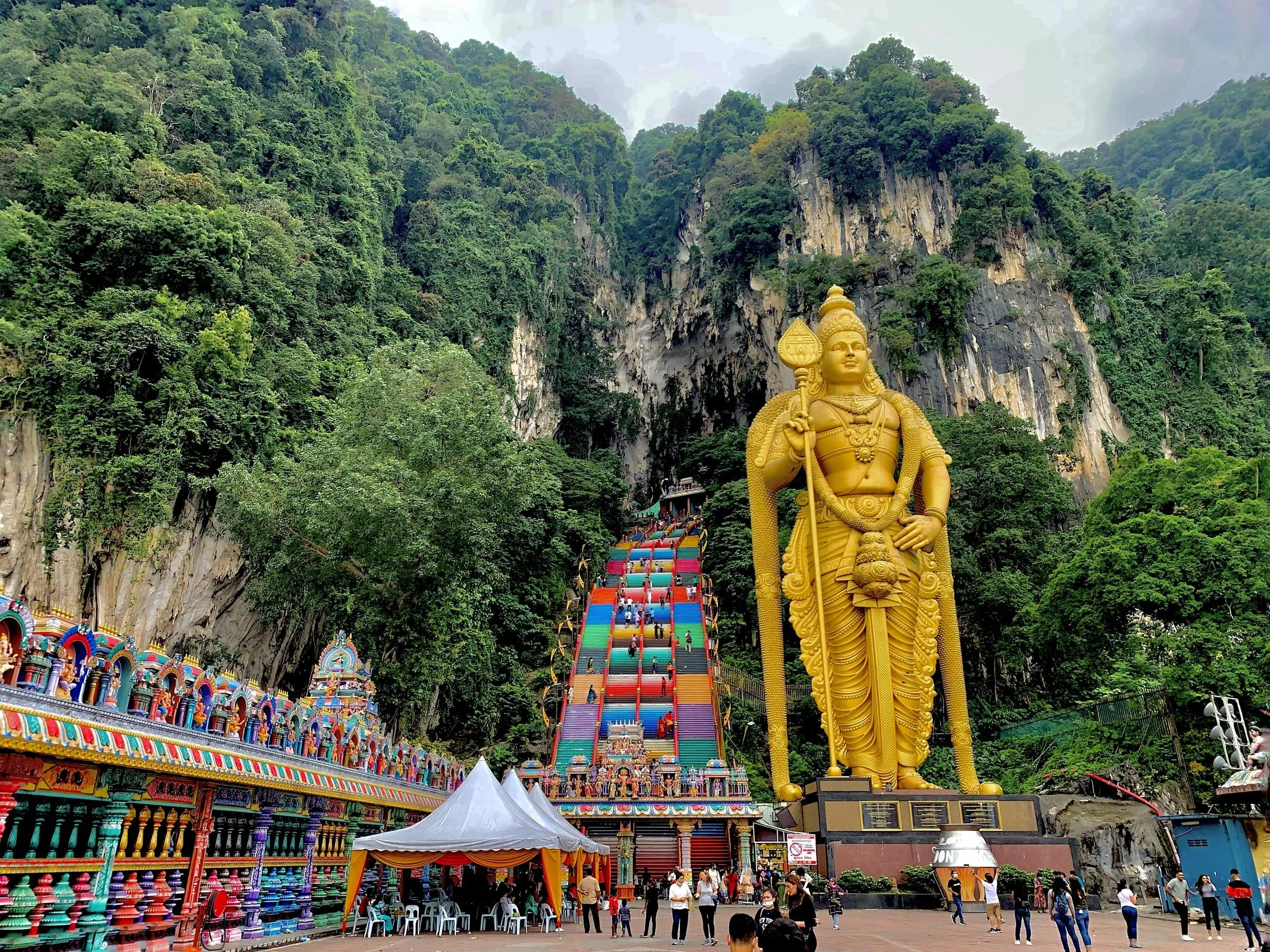 Estátua dourada de Murugan ao lado da escadaria colorida até a entrada da caverna, entre falésias cobertas de mata.