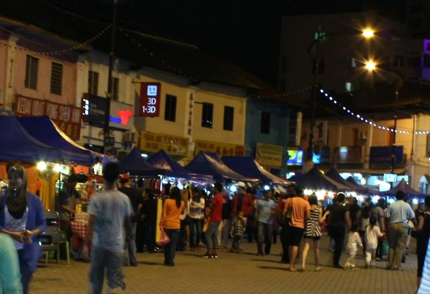 Barracas de roupas e visitantes no Bazaar Karat de Johor Bahru à noite, diante de fachadas iluminadas.
