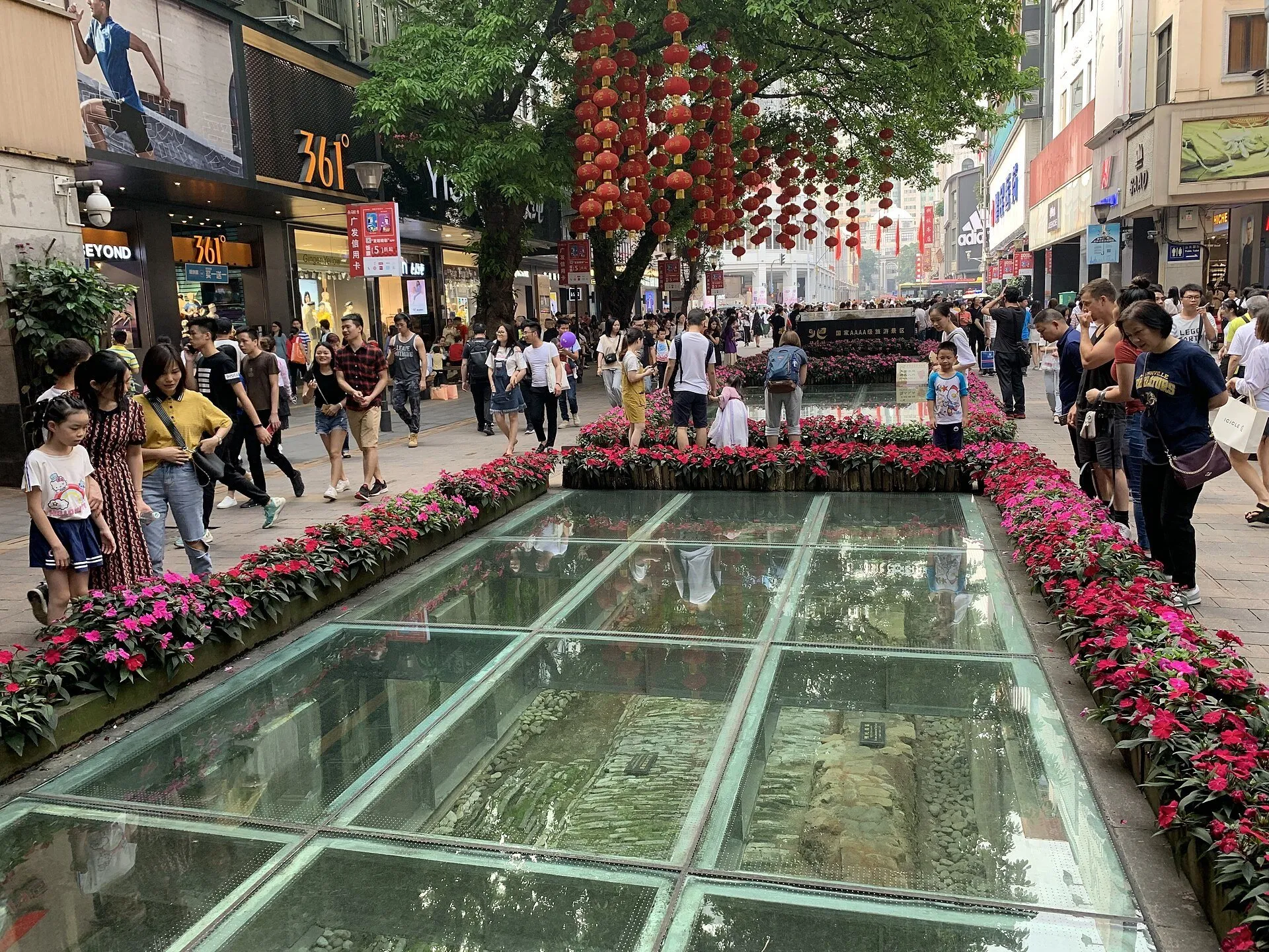 Pavimentos antigos sob vidro em Beijing Road, cercados por flores, lojas, lanternas vermelhas e pedestres.