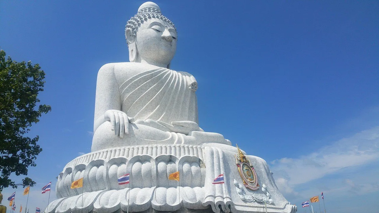 Estátua sentada do Big Buddha de Phuket, de mármore branco, sobre base em flor de lótus com bandeirolas, contra o céu azul.