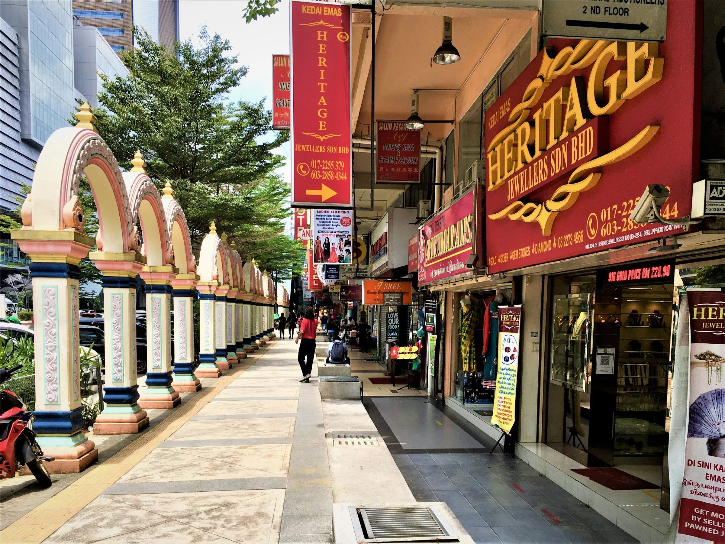 Calçada de Brickfields com arcos decorados em rosa, azul e dourado, ao lado de lojas de joias e tecidos, sob árvores.