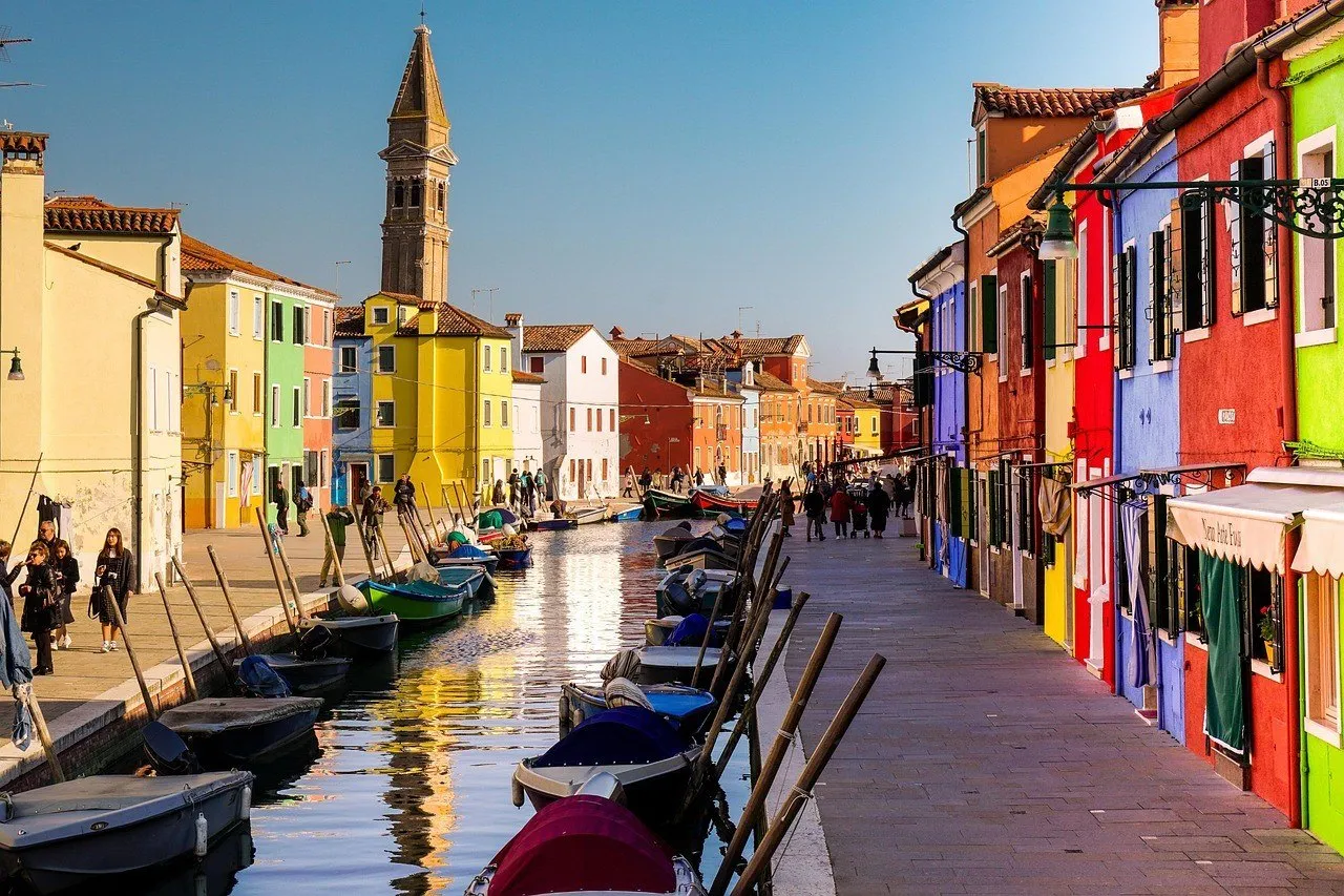 Casas coloridas de Burano junto ao canal, com barcos amarrados, reflexos e campanário ao fundo.