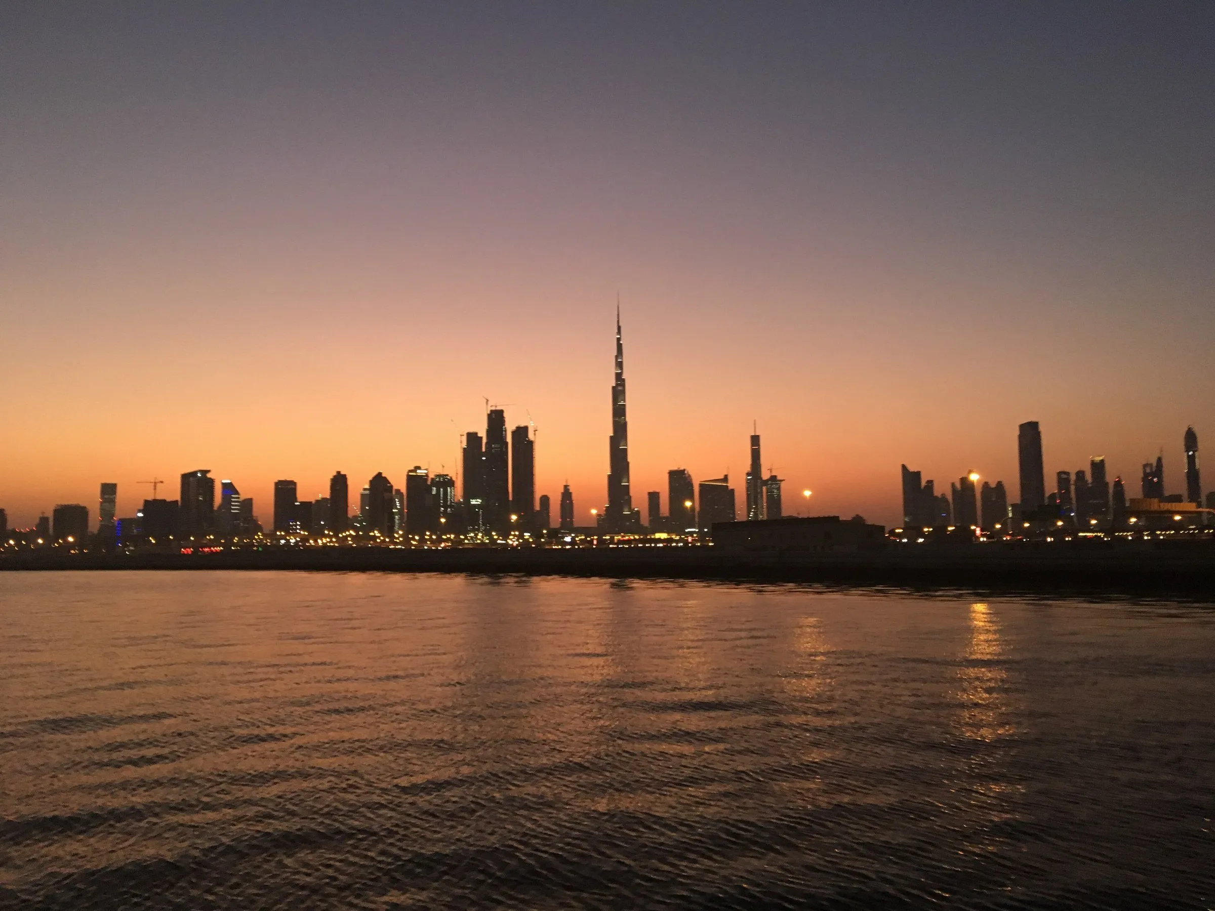 Skyline de Dubai com a silhueta do Burj Khalifa sobre o mar, sob um céu alaranjado e roxo logo após o pôr do sol.