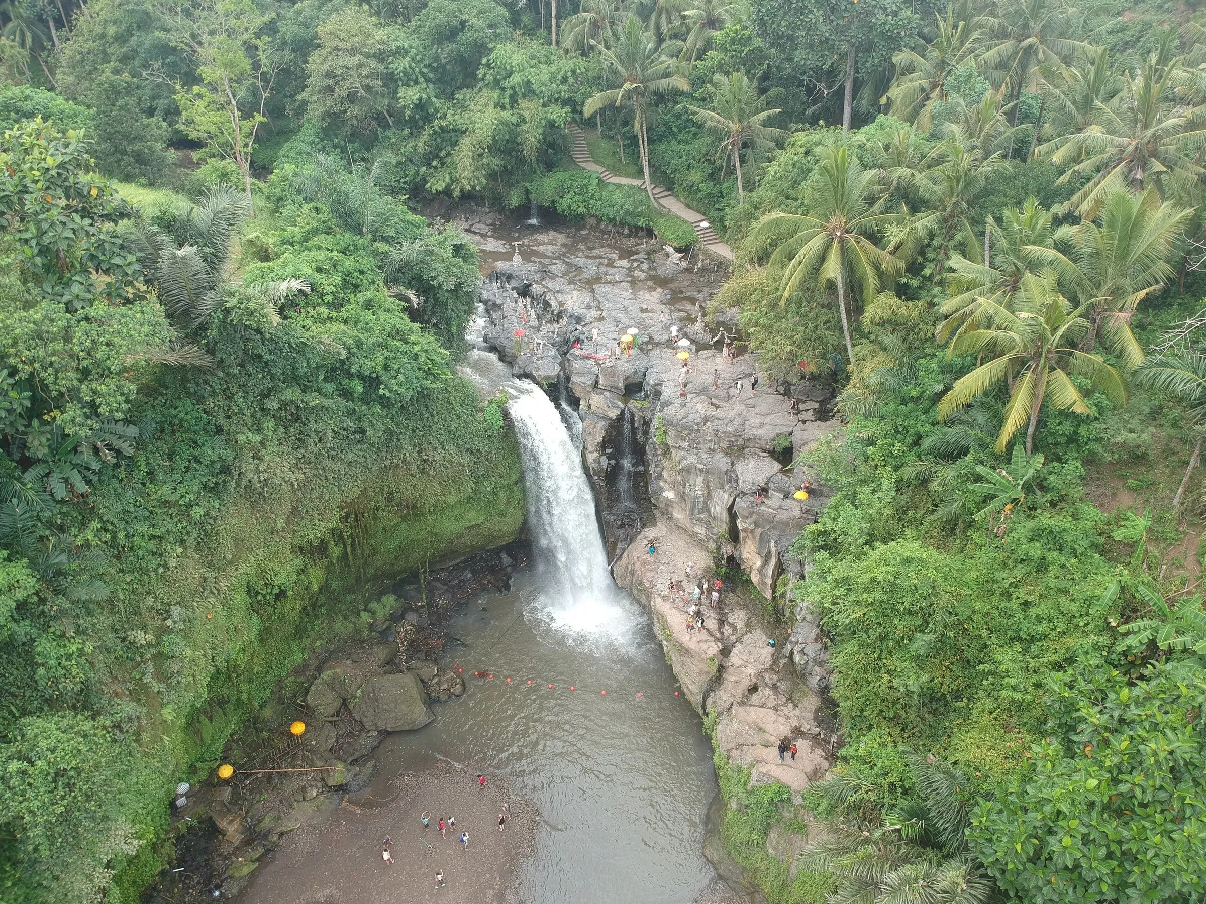 Cachoeira de Tegenungan vista do alto, cercada por palmeiras e vegetação, com visitantes pequenos junto às rochas.