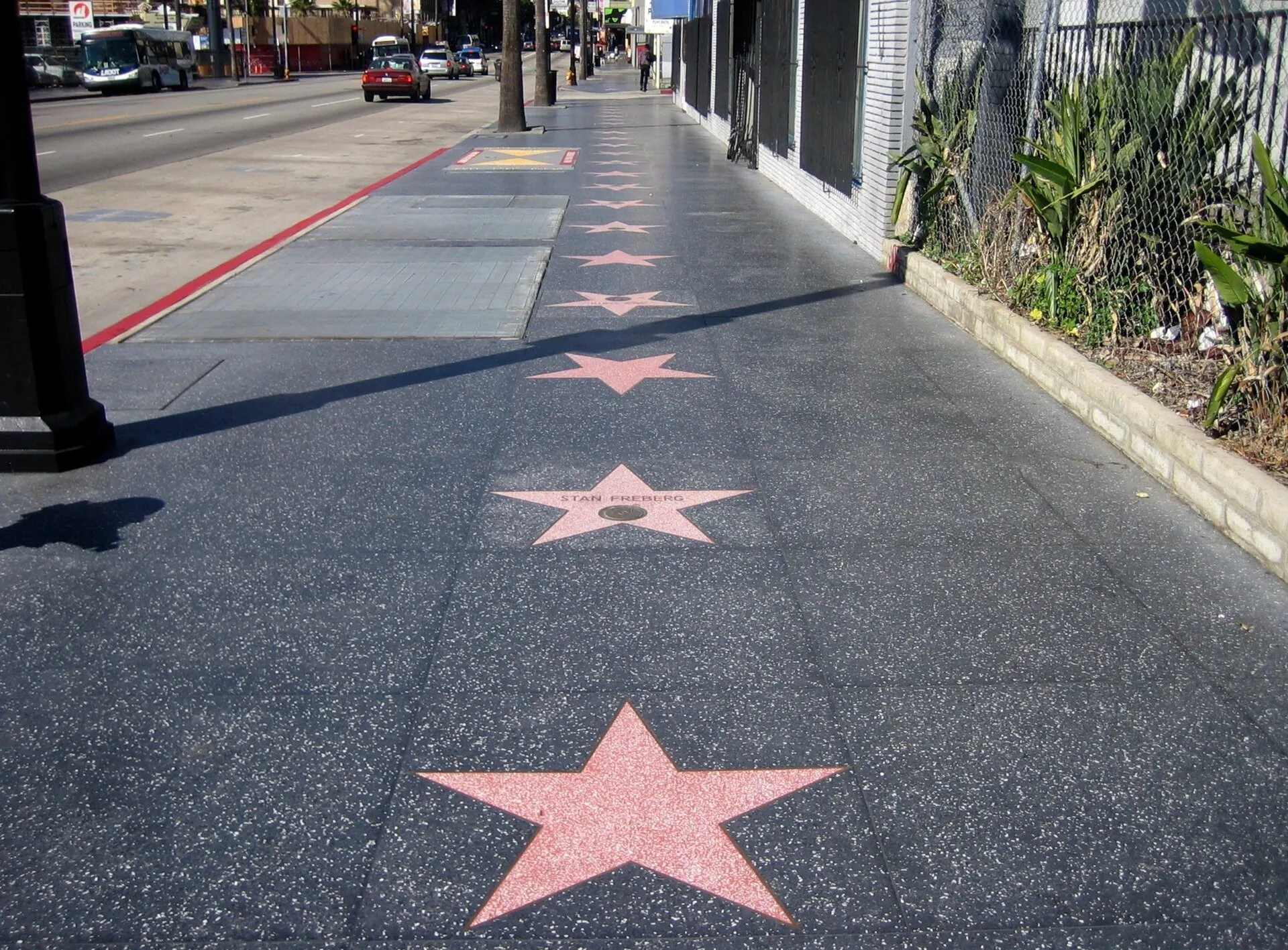 Estrelas da Calçada da Fama em sequência, ao lado de avenida e fachadas, em Hollywood.