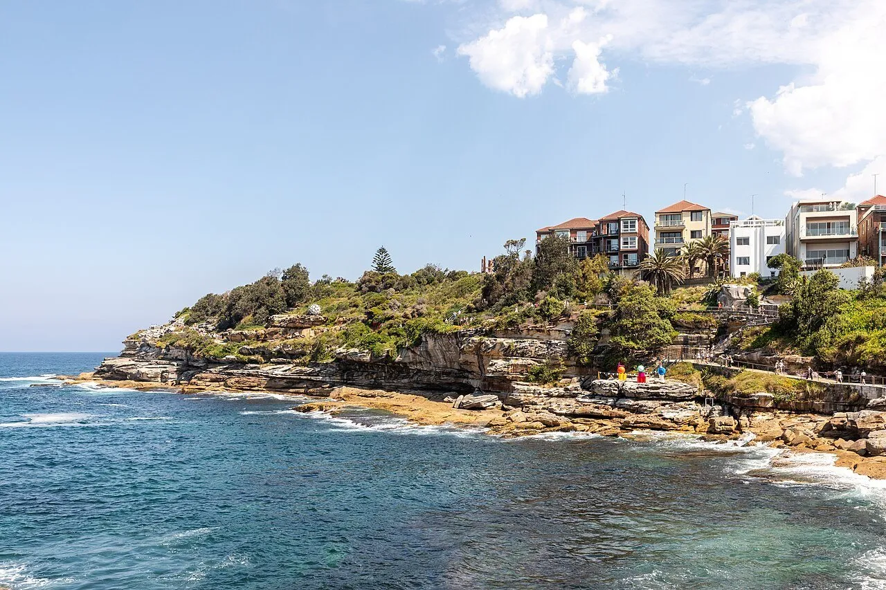 Trecho da caminhada Bondi–Bronte junto a rochas e mar azul, com guarda-corpo e casas na encosta.