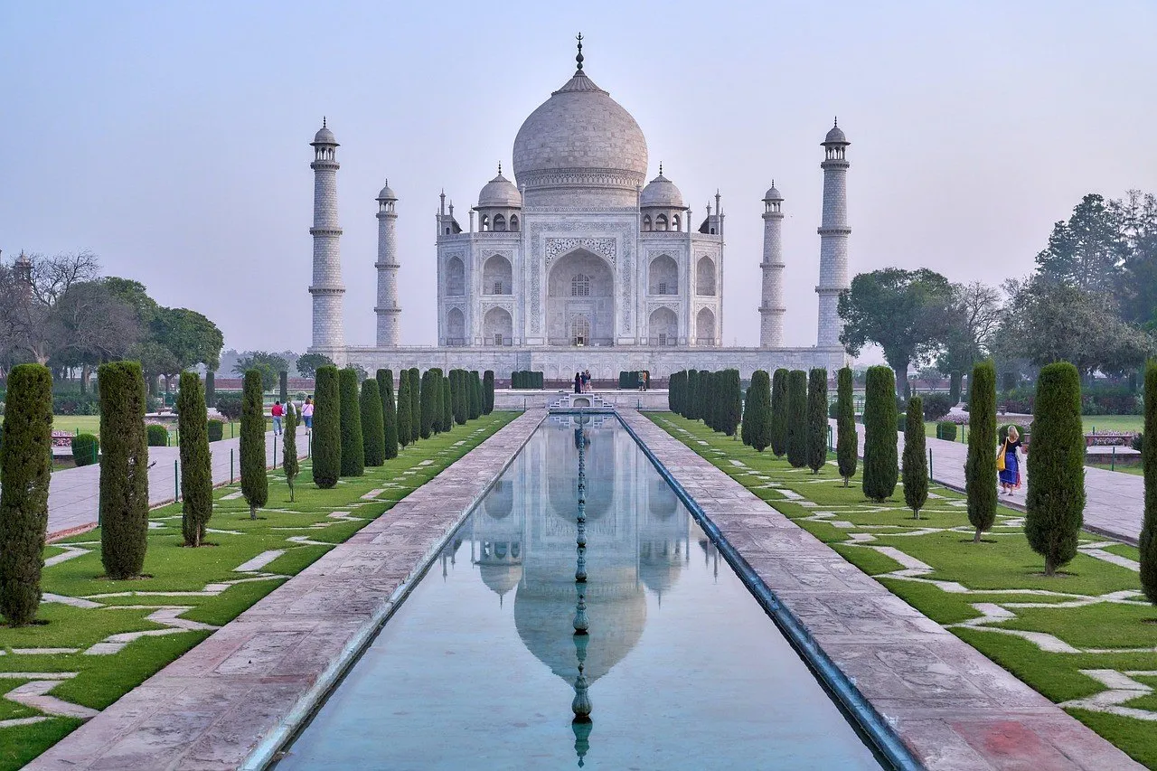 Taj Mahal refletido no espelho d'água central, entre ciprestes e caminhos do jardim.