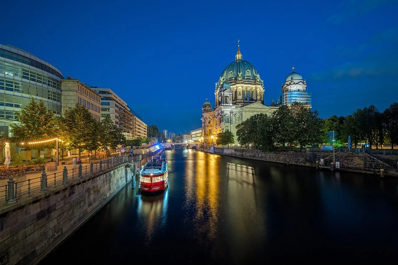 Catedral de Berlim iluminada junto ao Spree, com barco vermelho e reflexos dourados na água.