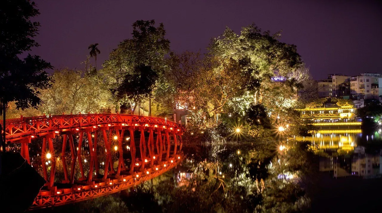 Ponte vermelha The Huc iluminada à noite, com árvores e reflexos no lago Hoan Kiem.