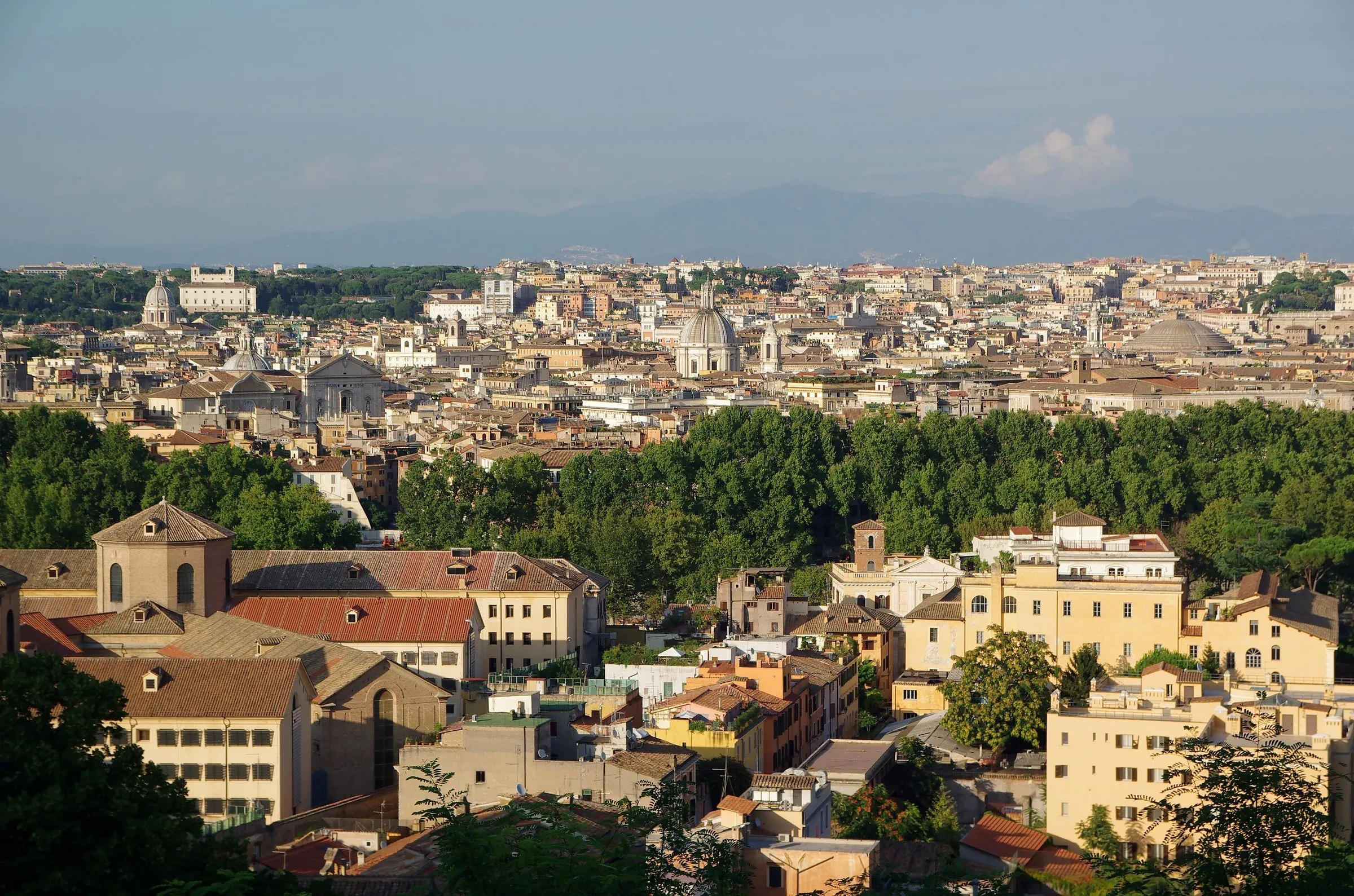 Panorama de Roma visto do alto, com telhados, cúpulas de igrejas e árvores sob luz suave de fim de tarde.