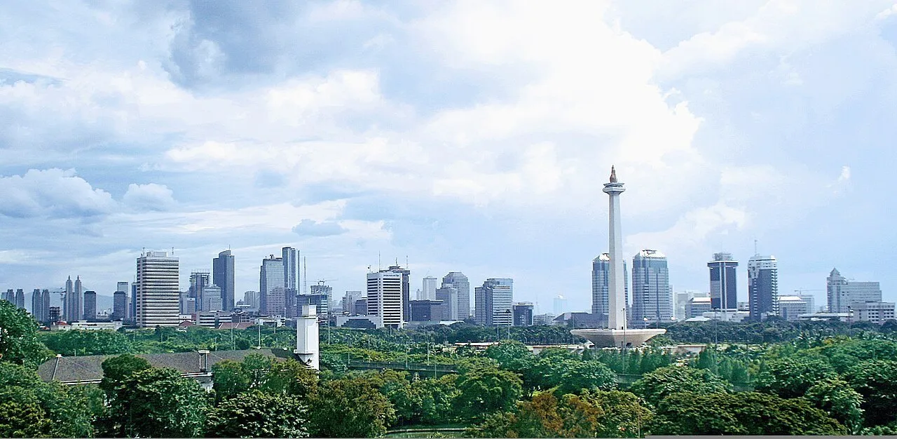 Panorama do Monas entre árvores, com edifícios de Jacarta ao fundo e céu com nuvens.