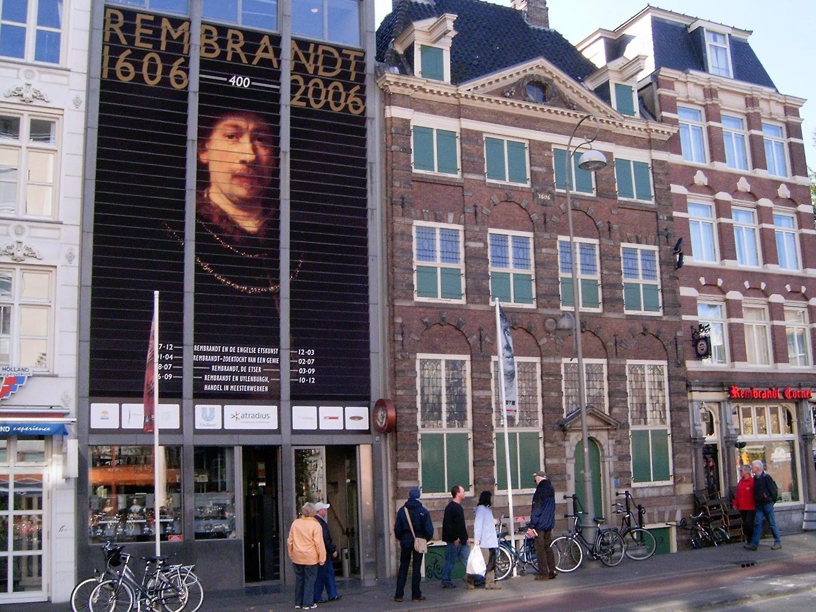 Fachada de tijolos e janelas verdes da Casa de Rembrandt ao lado da ampliação, com bicicletas e pedestres.