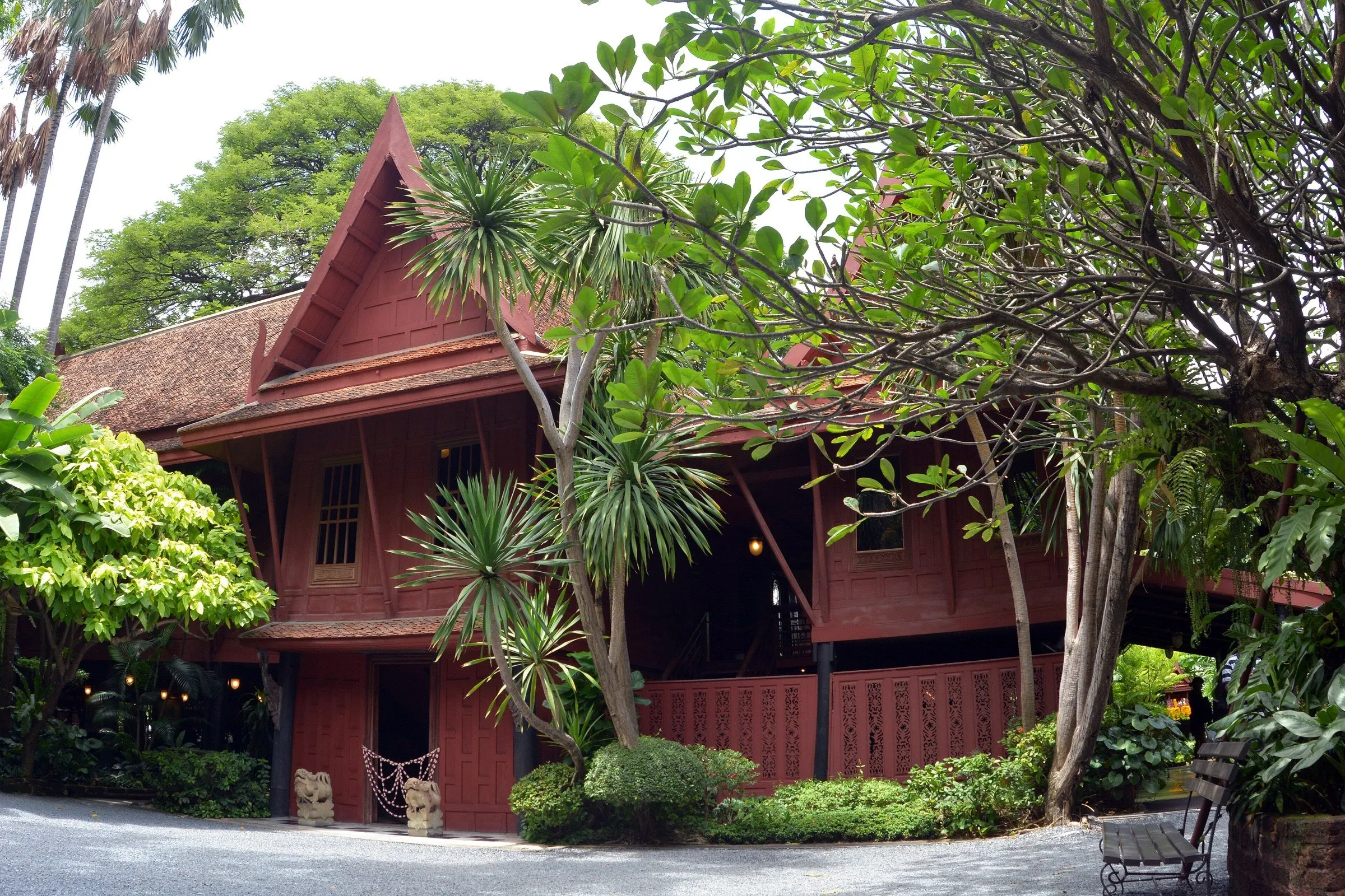 Casa tradicional tailandesa de madeira vermelha, a Casa de Jim Thompson, cercada de árvores e palmeiras.