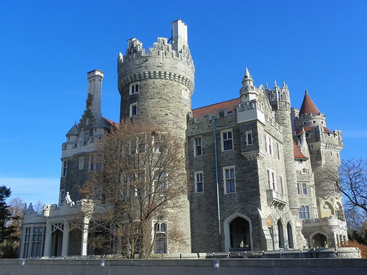Fachada de pedra de Casa Loma, com torres, telhados vermelhos e árvores sem folhas sob céu azul.
