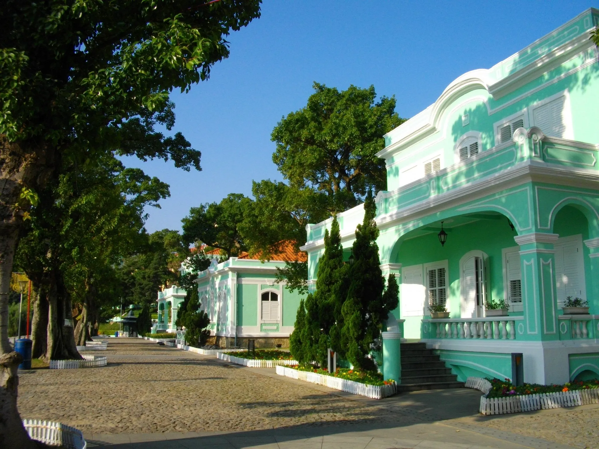 Casa-museu verde-água de dois andares com varanda, ao lado de um caminho de pedra sob árvores e céu azul.