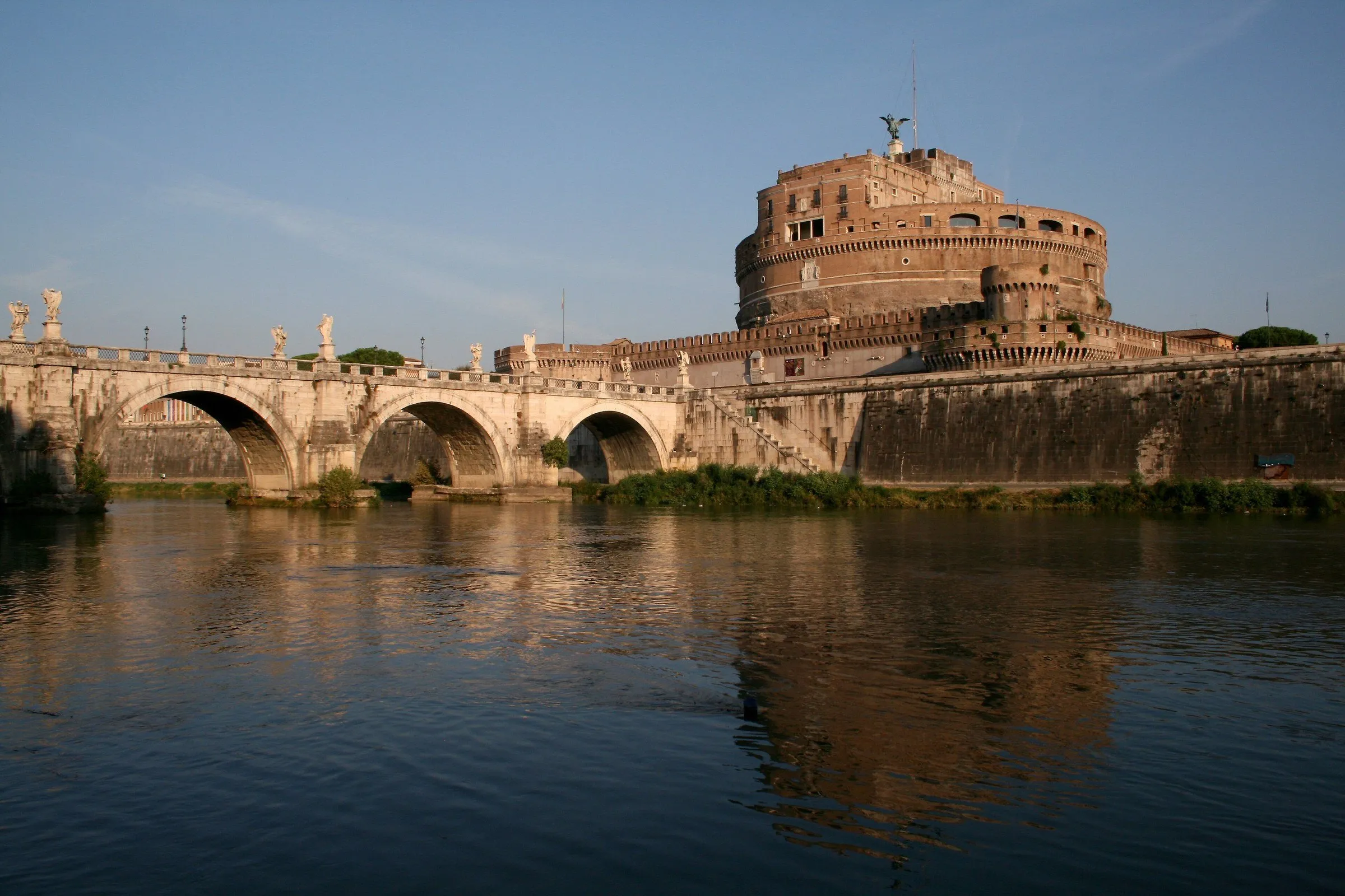 O Castel Sant'Angelo e a ponte de arcos de pedra sobre o rio Tibre, com reflexo na água sob luz dourada.