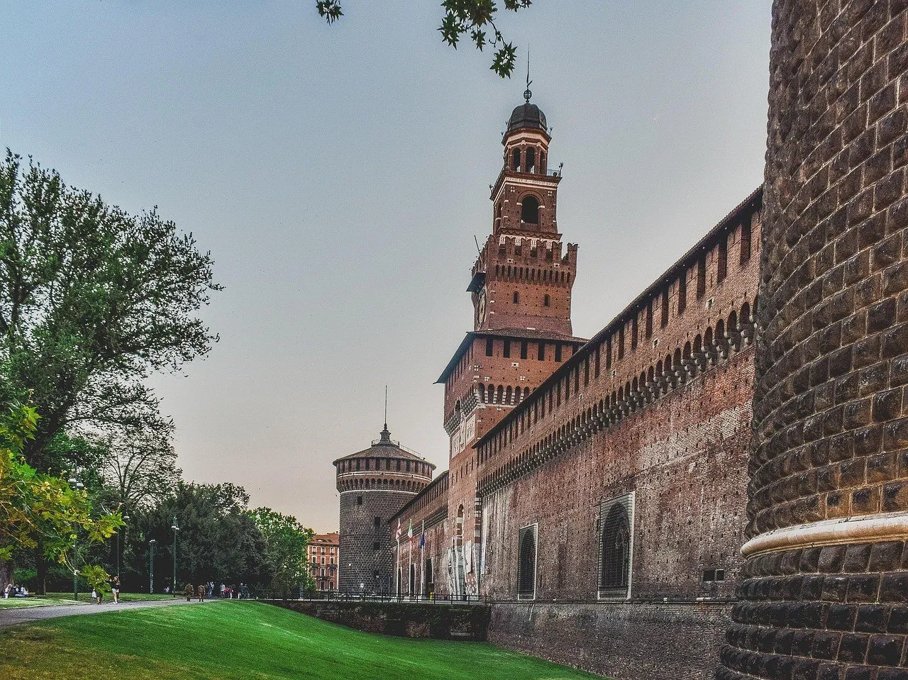 Muralhas de tijolos do Castello Sforzesco em perspectiva, com a torre do Filarete e gramado lateral.