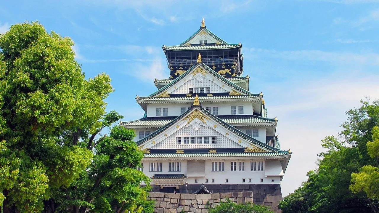 Torre branca do Castelo de Osaka com telhados verdes e detalhes dourados, entre árvores sob céu azul.