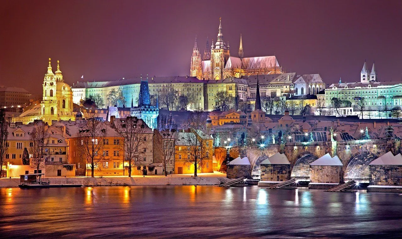 Castelo de Praga e Catedral de São Vito iluminados à noite, com telhados nevados e reflexos no Vltava.