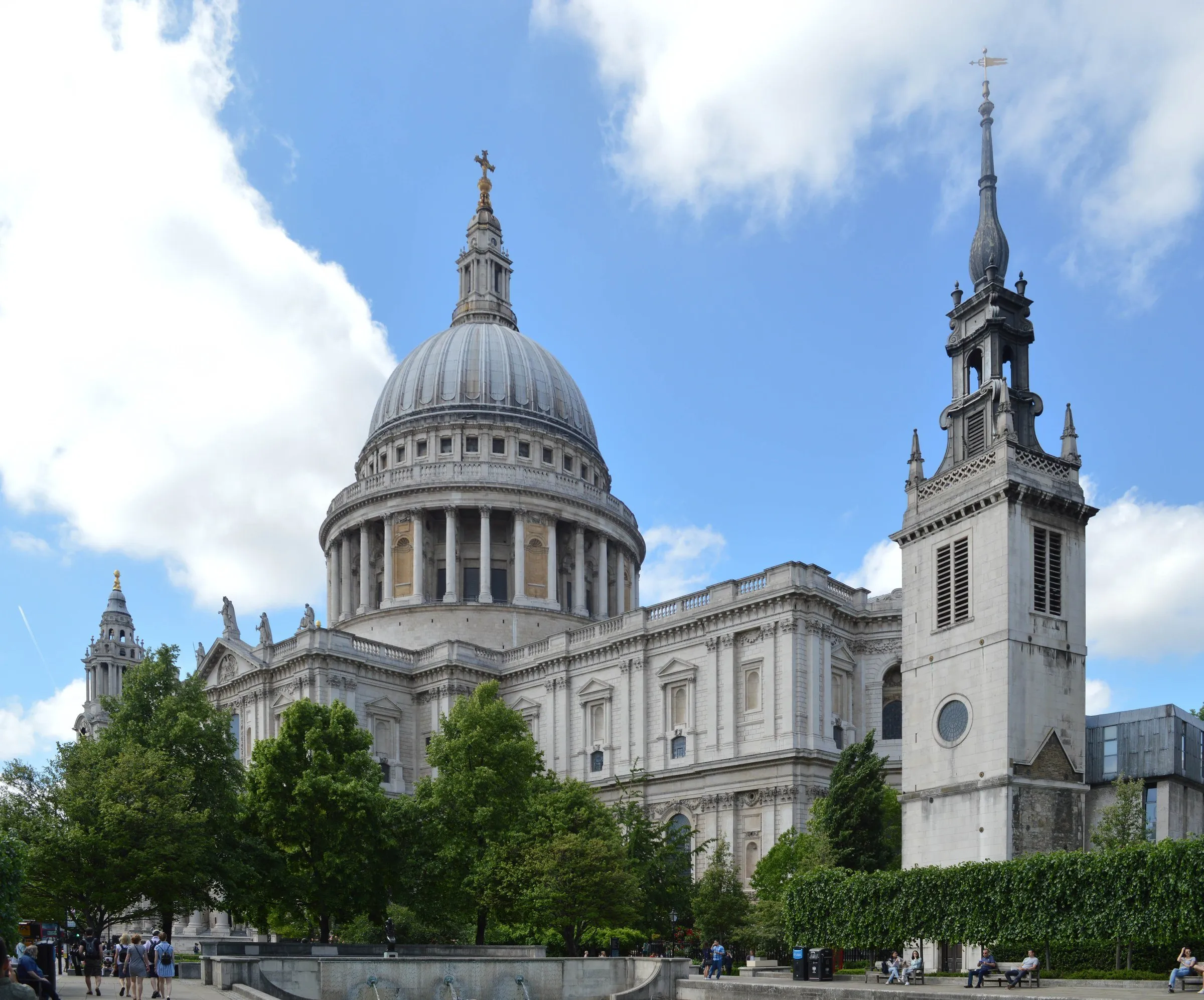 Catedral de St Paul com a grande cúpula e uma torre ao lado, vistas por entre árvores sob céu azul com nuvens.