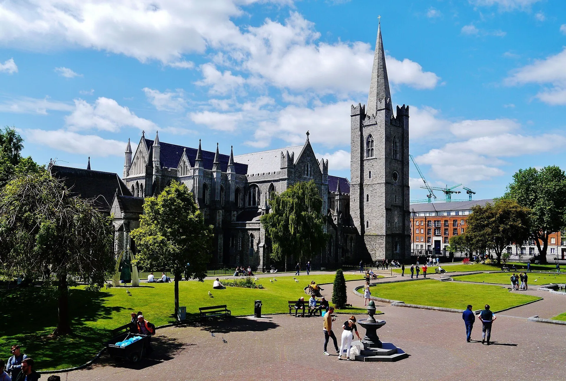 Catedral de St Patrick com torre pontiaguda, jardins e visitantes nos caminhos, sob céu azul com nuvens.