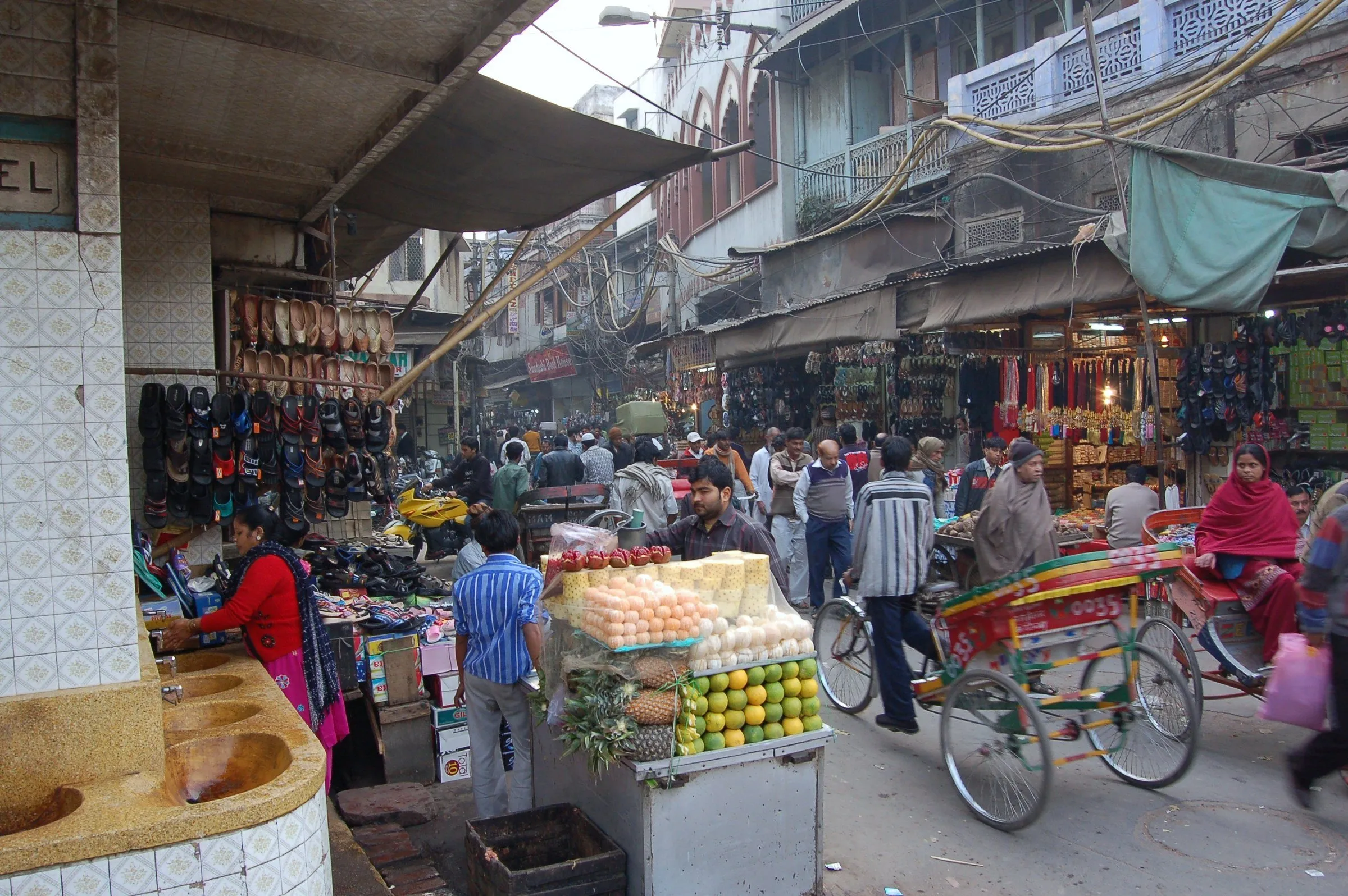 Rua movimentada do mercado de Chandni Chowk, com bancas de sapatos e frutas, um riquixá colorido e gente entre toldos e fios.