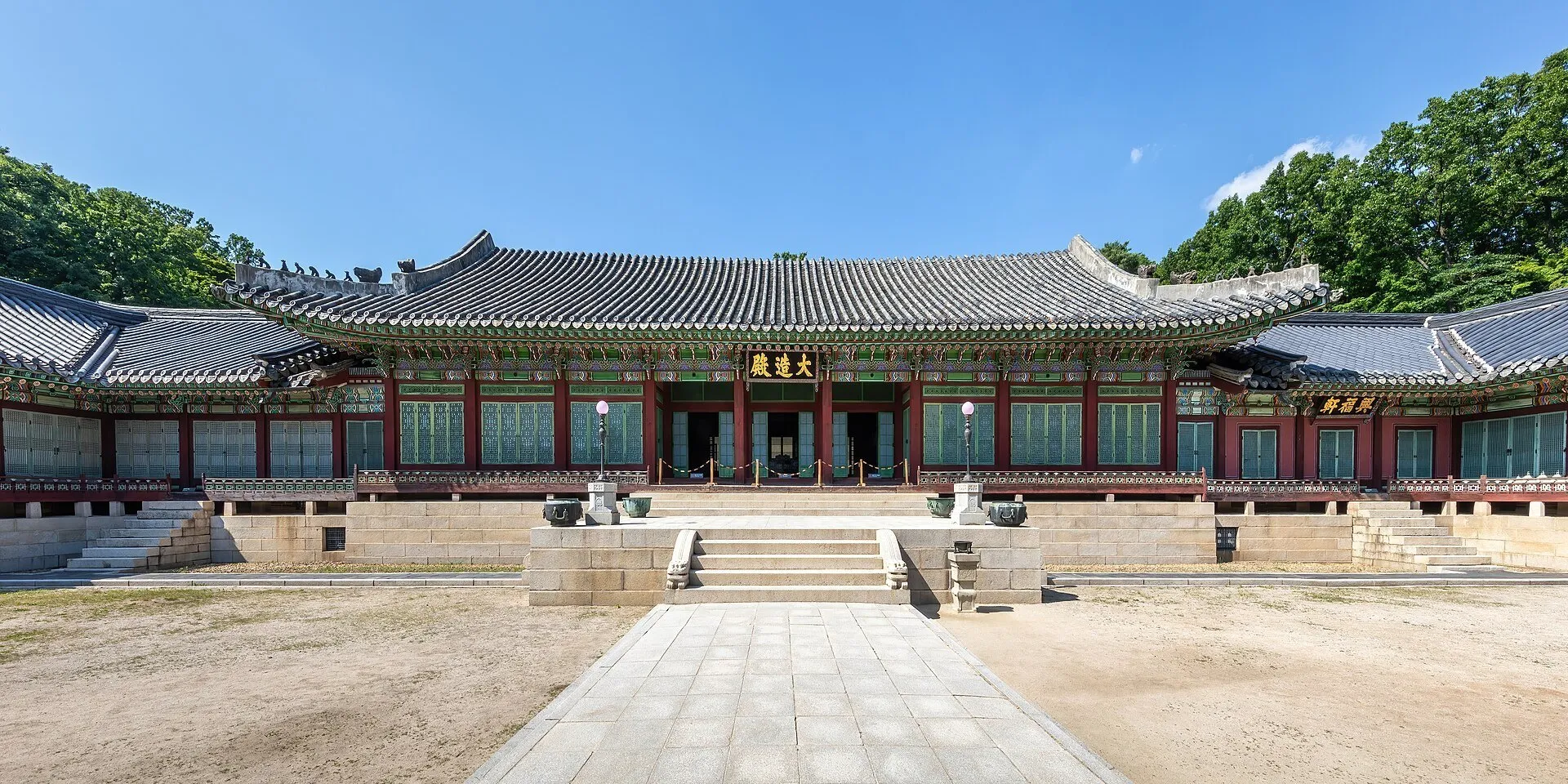 Fachada de um pavilhão de Changdeokgung com telhado curvo, pátio e árvores ao fundo.