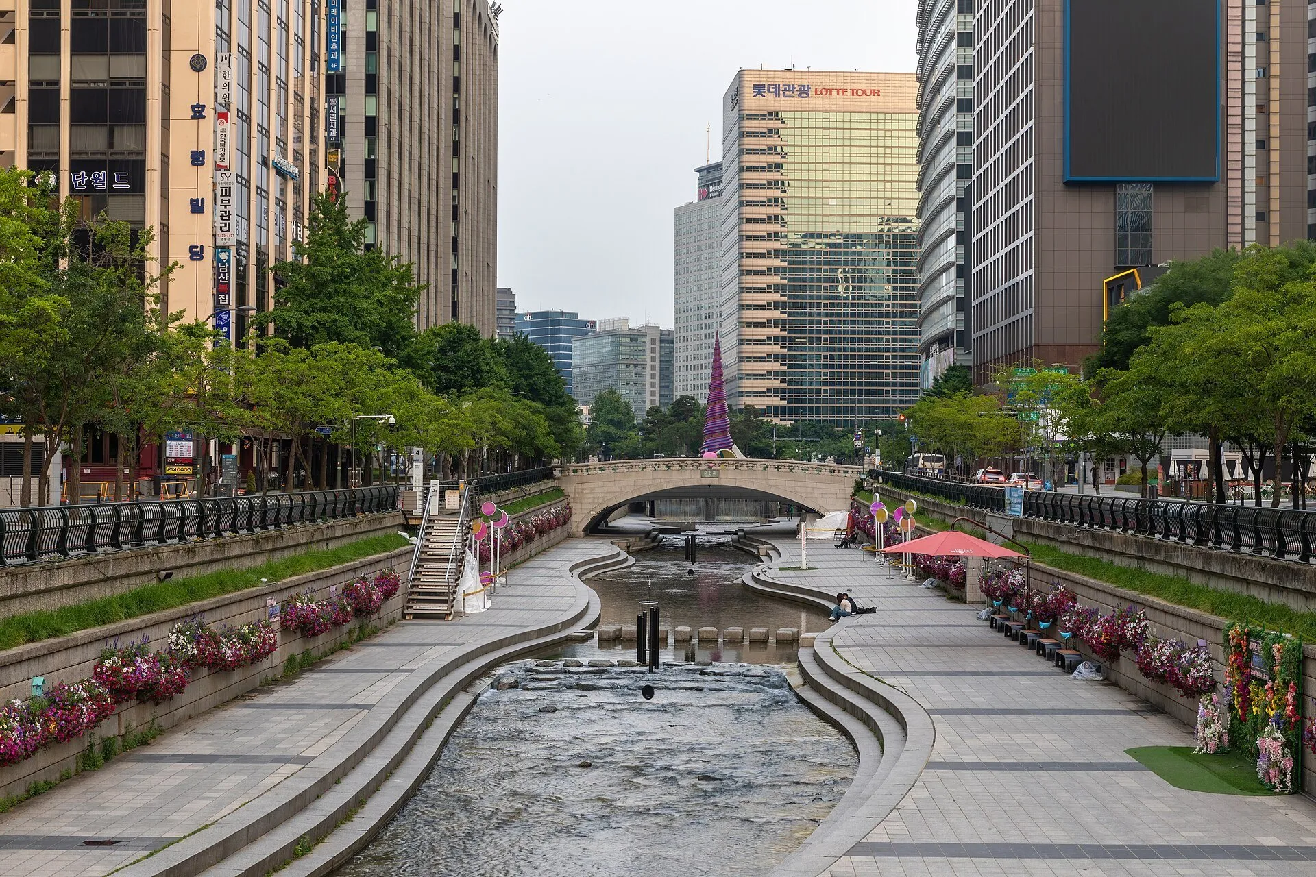 Córrego Cheonggyecheon entre passeios com flores, uma ponte de pedra e edifícios de Seul.