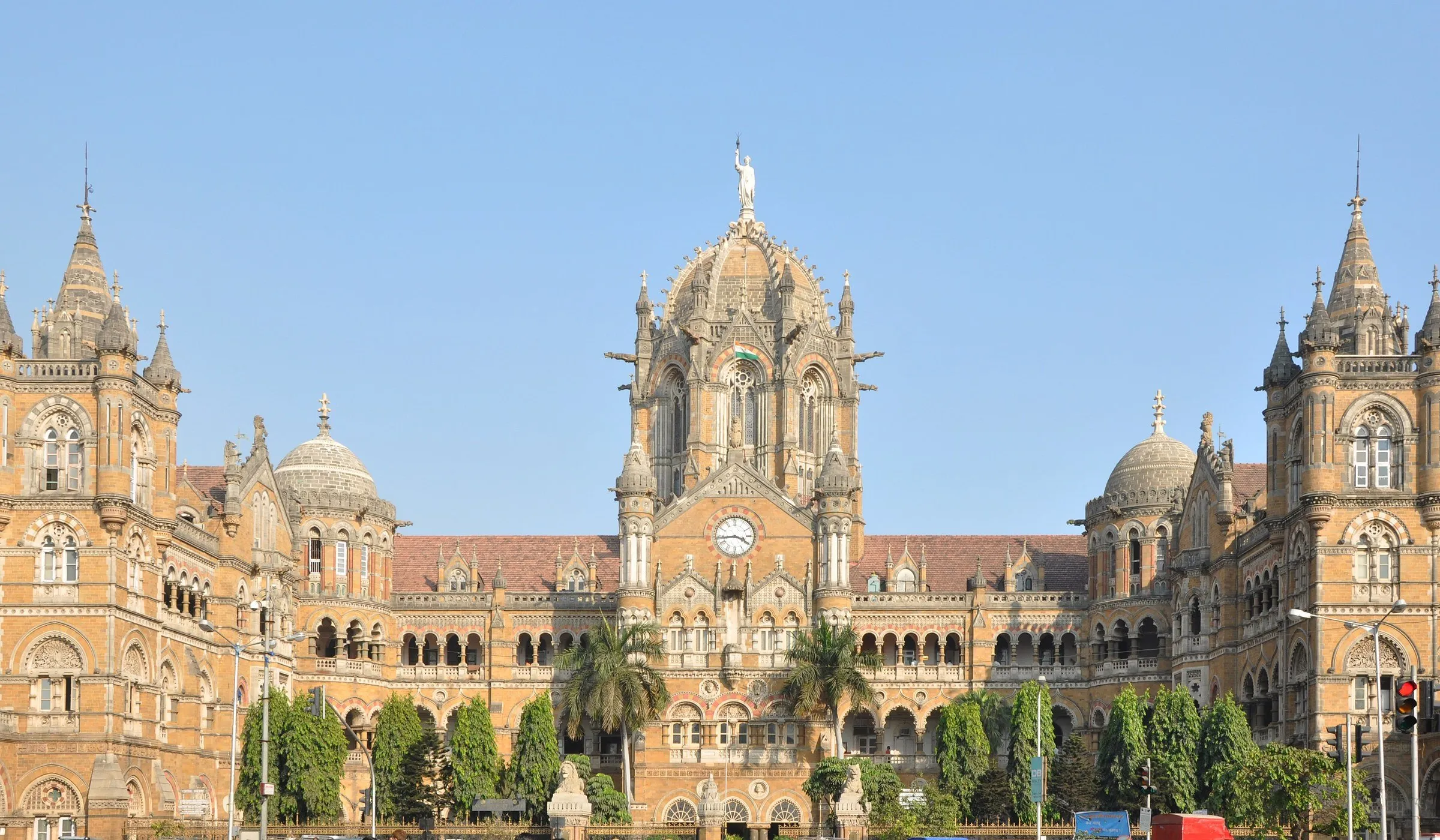Fachada central da estação Chhatrapati Shivaji Maharaj Terminus, em Mumbai, com cúpula e relógio sob céu azul sem nuvens.