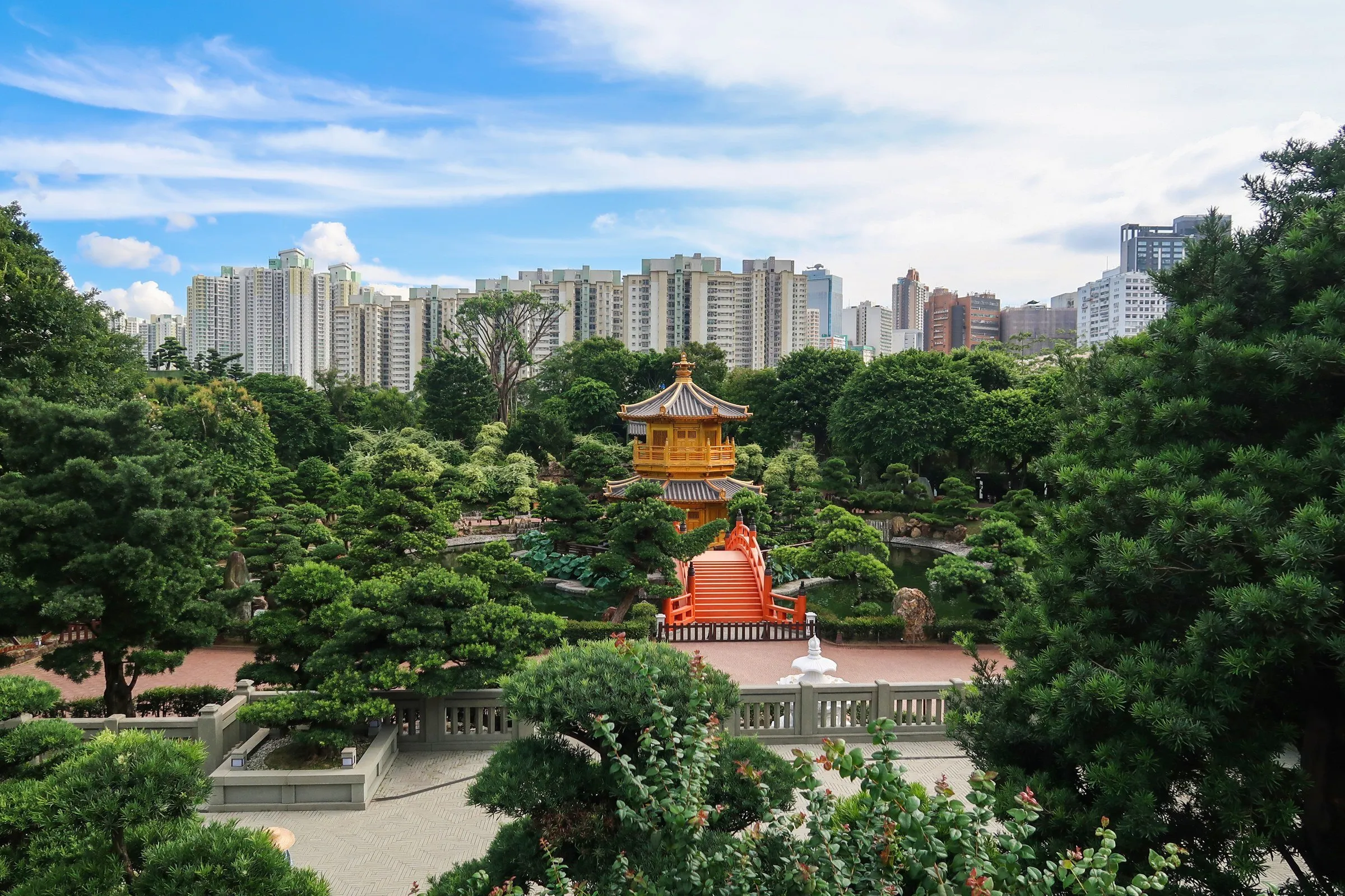 Pavilhão dourado de telhado oriental sobre ponte laranja, entre pinheiros podados, com prédios ao fundo sob céu azul.