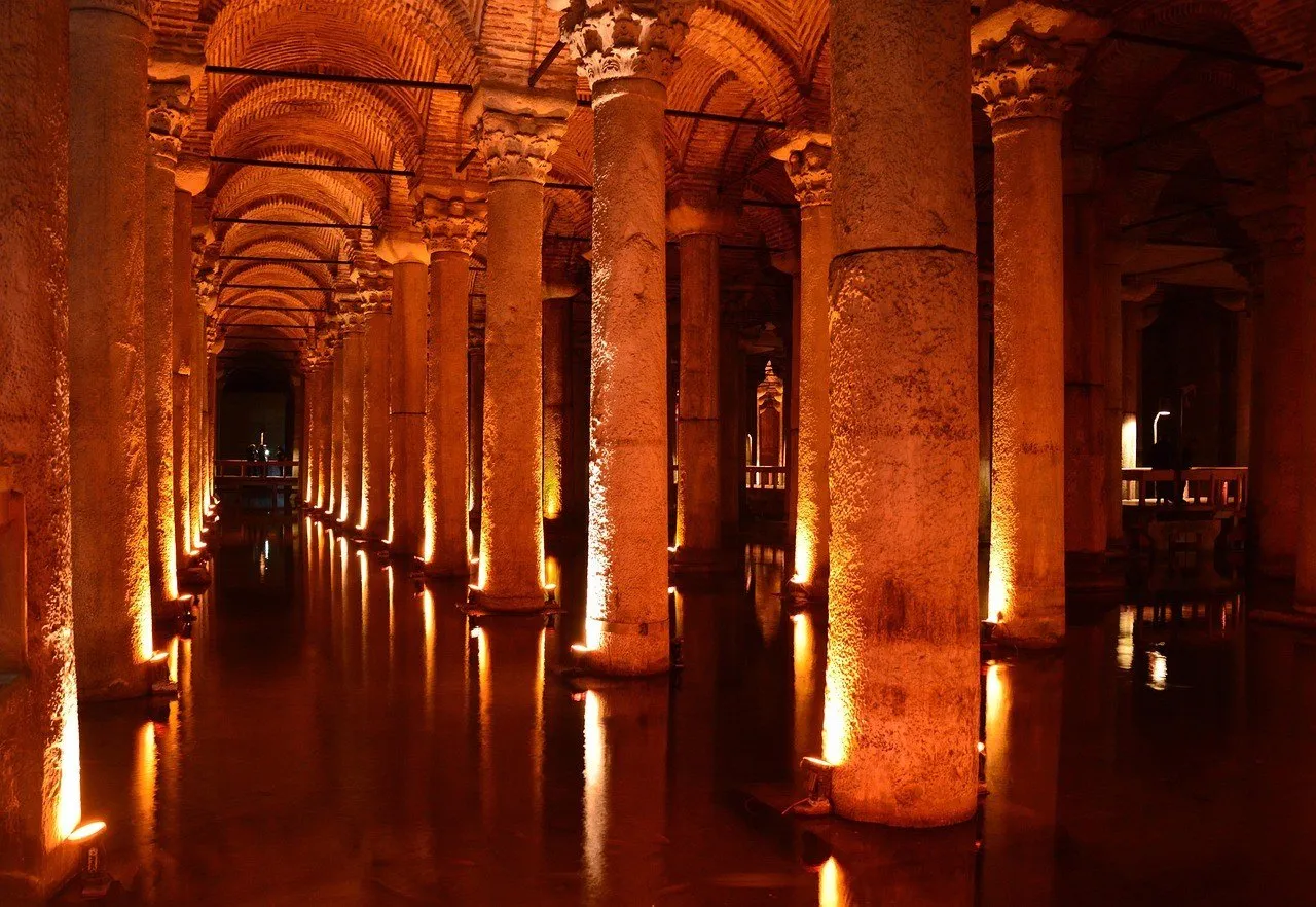 Corredor de colunas antigas com capitéis iluminados em laranja, abóbadas de tijolo e reflexo na água da cisterna.