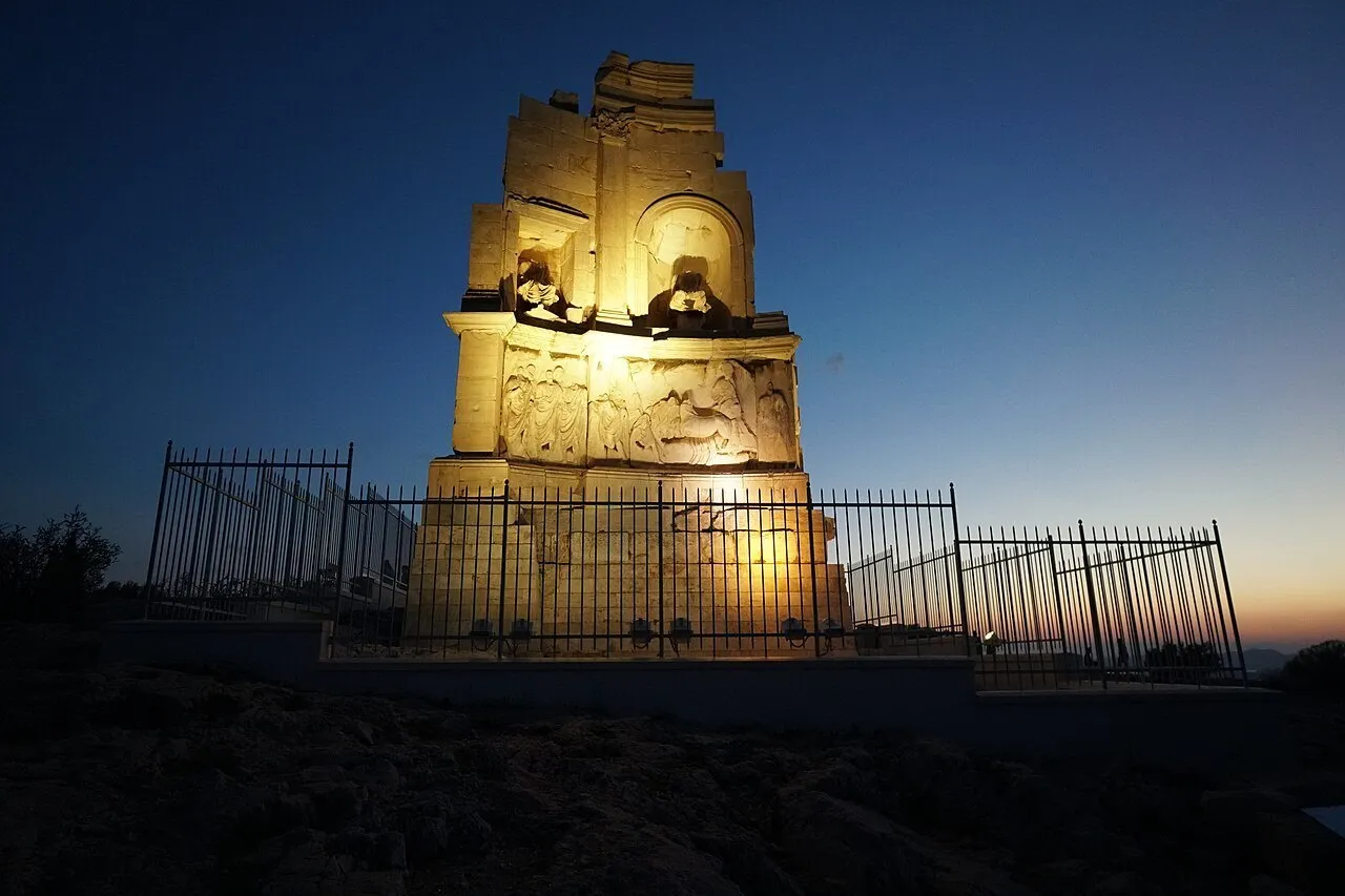 Monumento de Filopapo iluminado ao anoitecer, atrás de grade de proteção, sob céu azul-escuro em Atenas.