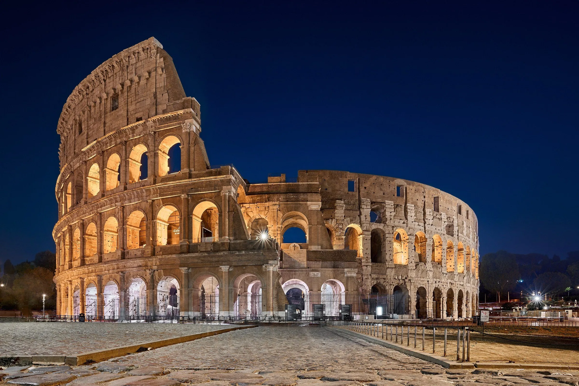 Fachada externa do Coliseu, em Roma, iluminada ao anoitecer sob um céu azul profundo, com a praça vazia à frente.