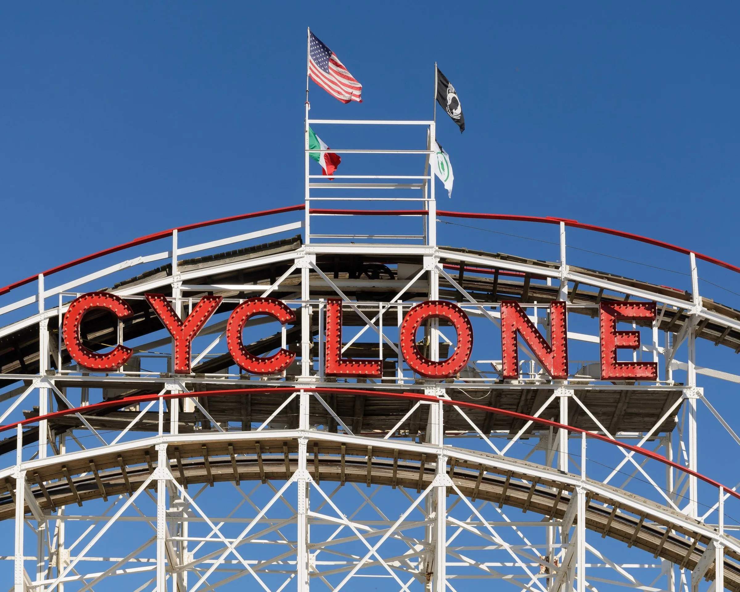 Letreiro vermelho CYCLONE no topo da montanha-russa de madeira de Coney Island, com bandeiras e céu azul ao fundo.