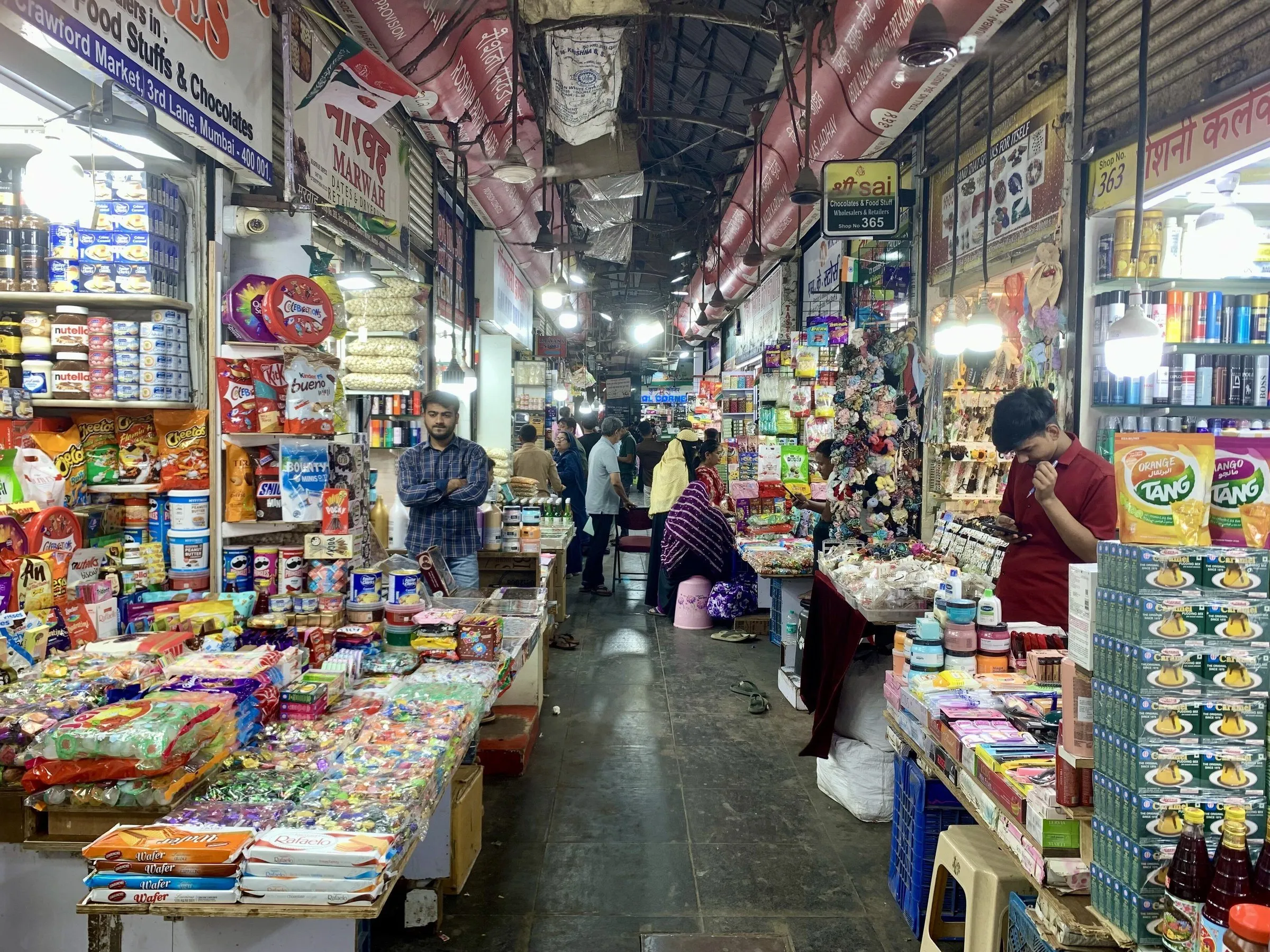 Corredor interno do Crawford Market, em Mumbai, com bancas lotadas de produtos, lâmpadas penduradas e clientes ao fundo.