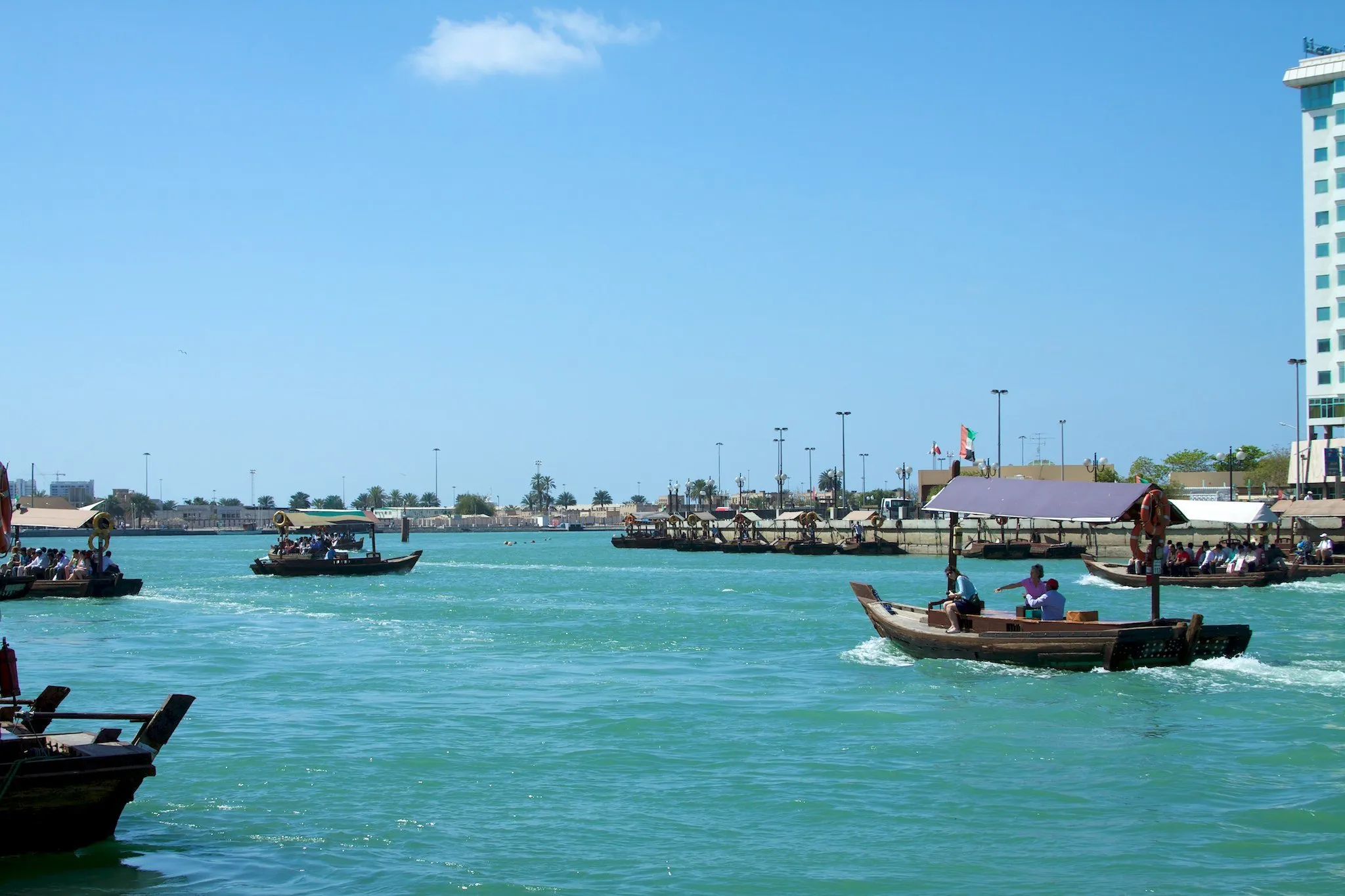 Abras de madeira com passageiros cruzando as águas turquesa do Dubai Creek sob céu azul, com a margem de Deira ao fundo.