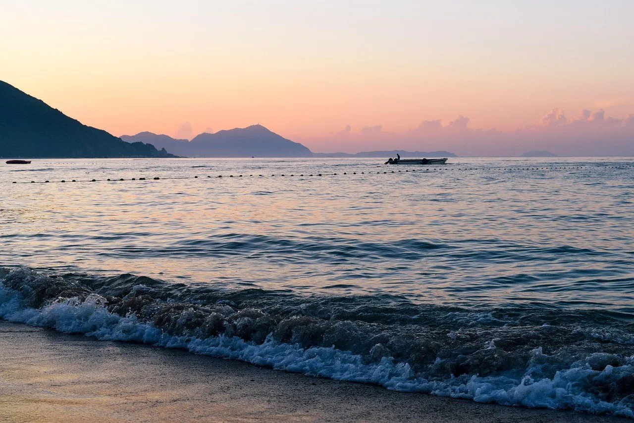 Ondas chegando à praia de Dameisha ao amanhecer, com linha de boias, um barco ao longe e morros contra o céu alaranjado.