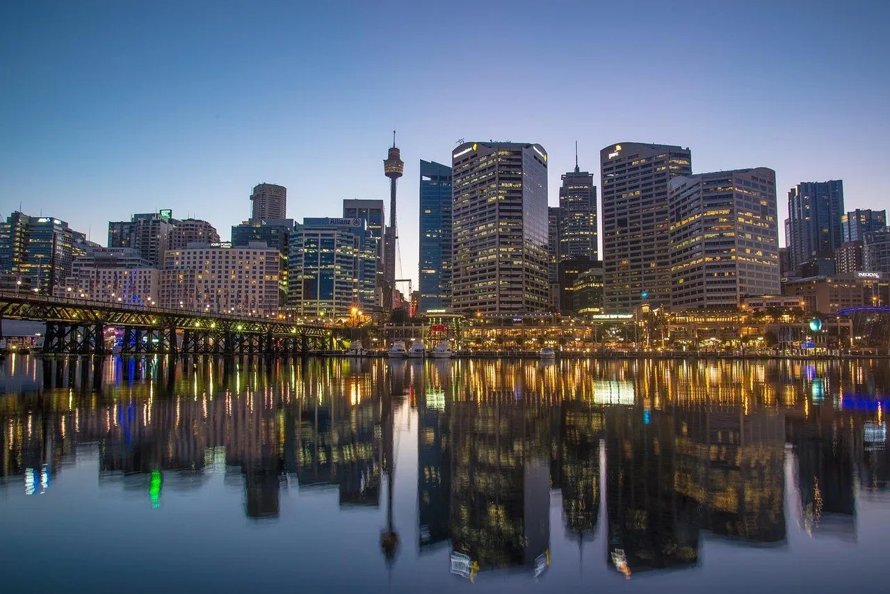 Edifícios iluminados e ponte de pedestres refletidos nas águas calmas de Darling Harbour sob céu azul de crepúsculo.