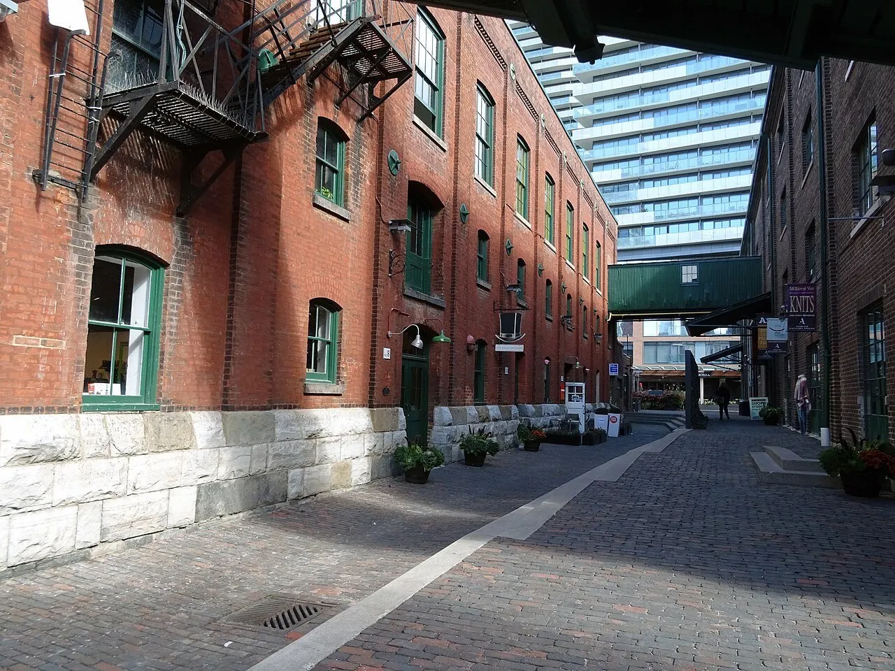 Passagem de pedestres no Distillery District entre edifícios de tijolos, com janelas verdes e passarela elevada.