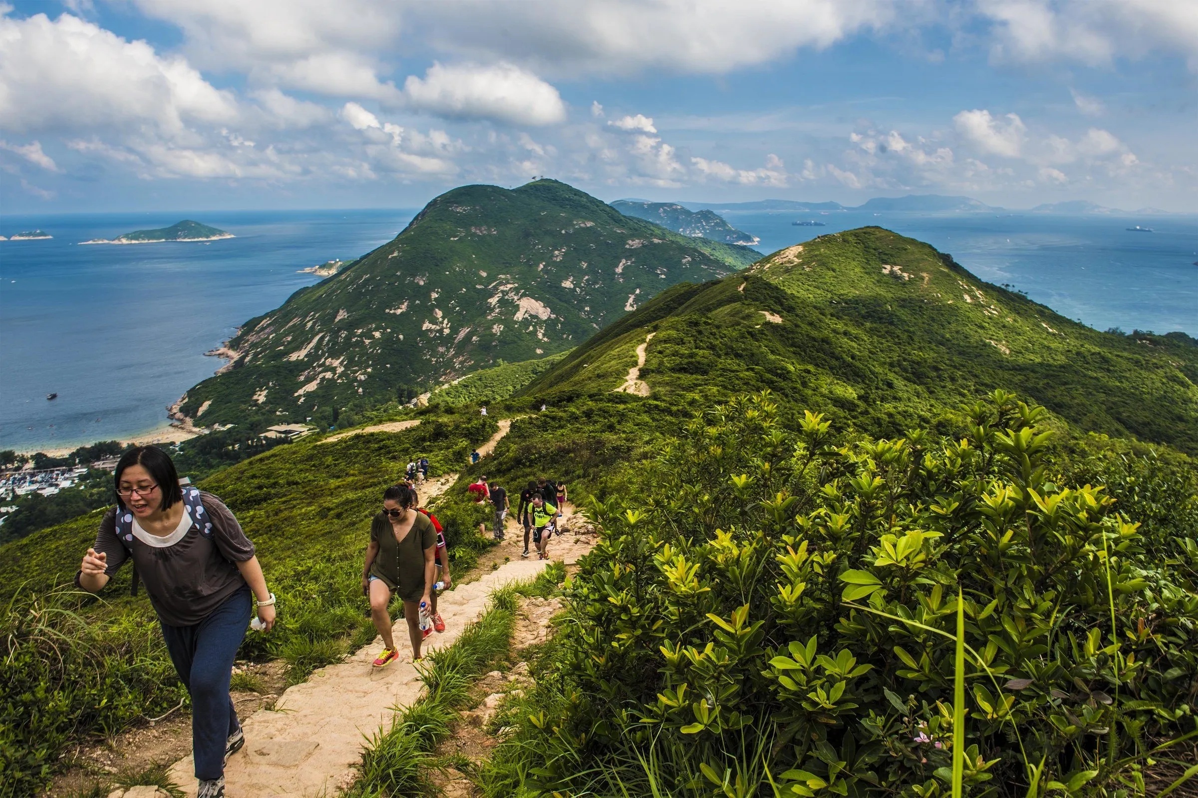Trilheiros subindo o caminho de terra na crista do Dragon's Back, com morros verdes, mar e ilhas ao fundo sob nuvens.