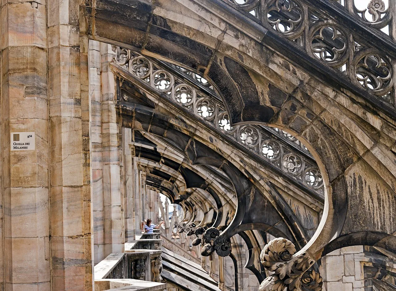 Arcobotantes de mármore e balaustradas nos terraços do Duomo de Milão, com visitantes ao fundo.